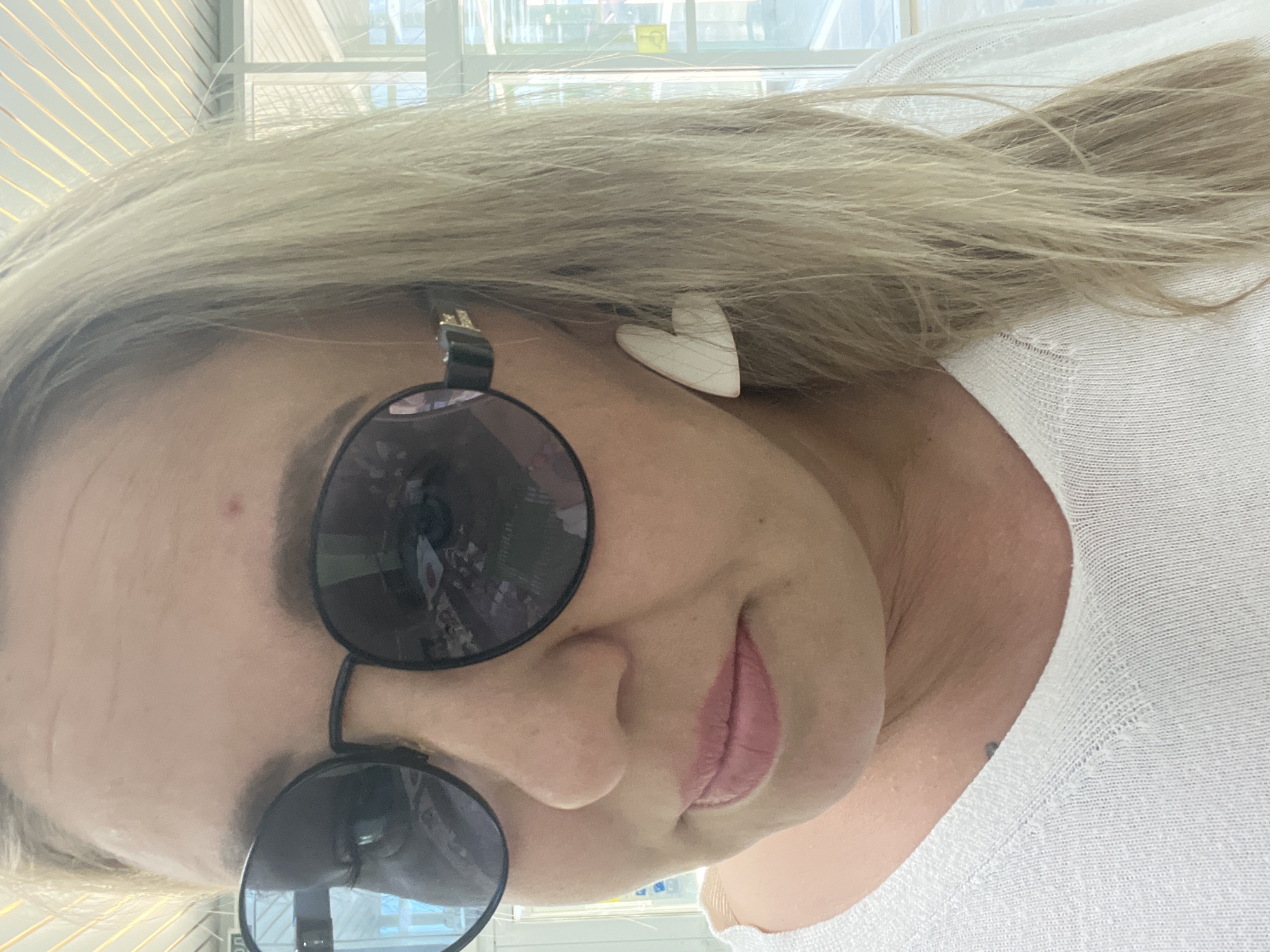 A woman with blonde hair is wearing round sunglasses and a white heart-shaped earring. She is wearing a white shirt. The reflection in her sunglasses shows a building with windows. The location is the city of Chaikovsky, Russia.