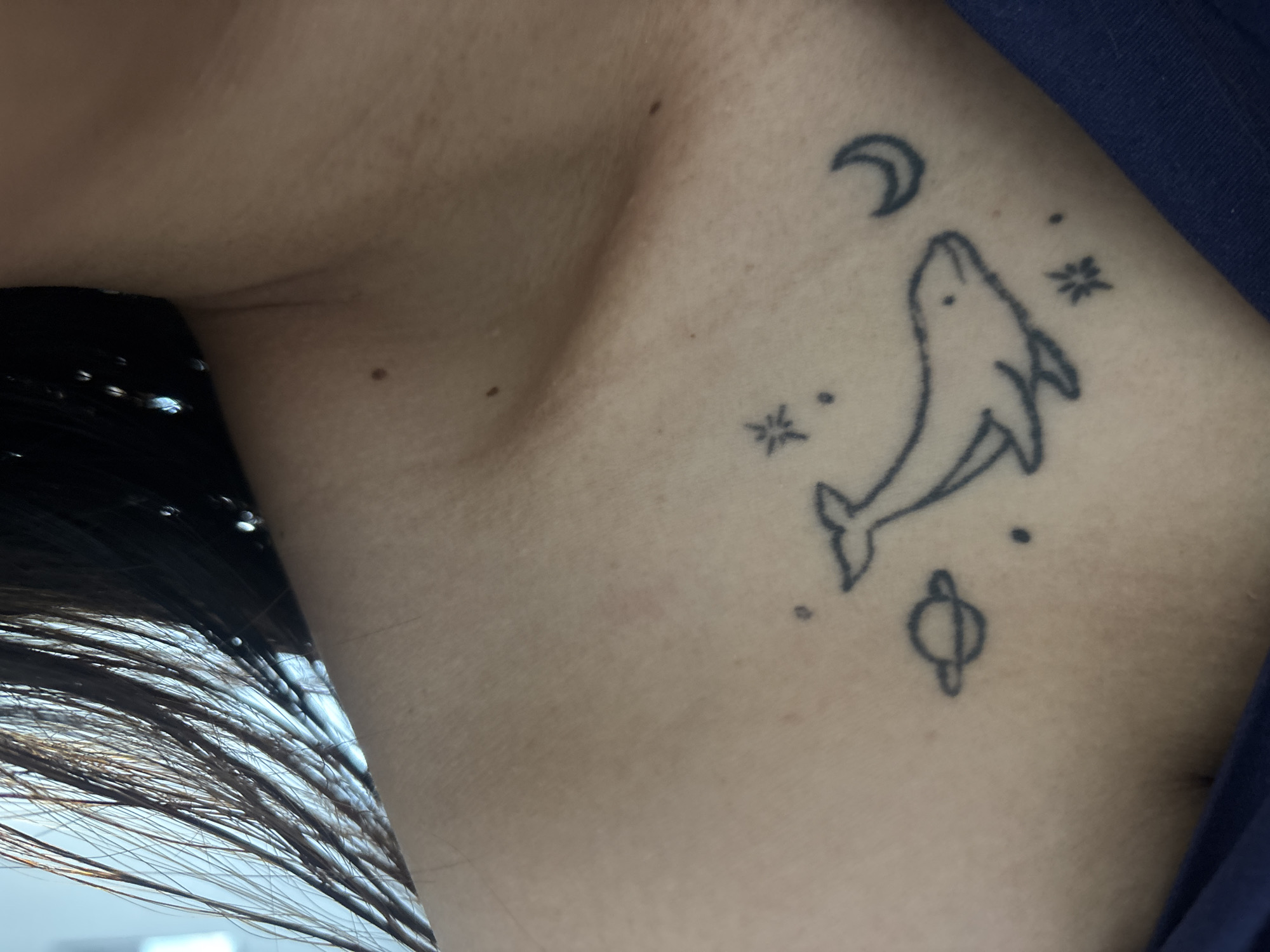 The image shows a close-up view of a person's upper chest and neck area, featuring a black line tattoo of a whale with a crescent moon, a ringed planet, and stars. The tattoo is located on the left side of the chest, below the collarbone. The person's skin is visible, along with strands of dark hair falling over the shoulder. The background is softly blurred, with a hint of blue fabric visible in the lower right corner. No specific location in Dubai, United Arab Emirates, can be determined from the image.