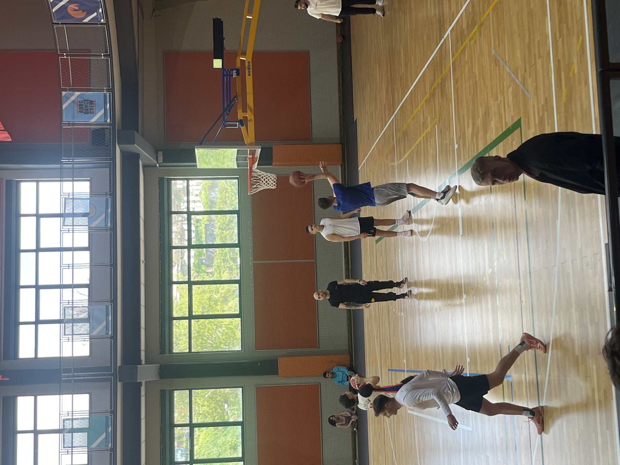 A basketball game is in progress at a gymnasium in Blagoevgrad, Bulgaria. Several people are participating or watching. One player in a blue shirt is shooting the ball. A tall basketball hoop with a net stands in front of a wall with square windows. The gymnasium has a wooden floor with lines and markings, and the walls have orange and red panels. There are posters or signs on the walls above the windows.