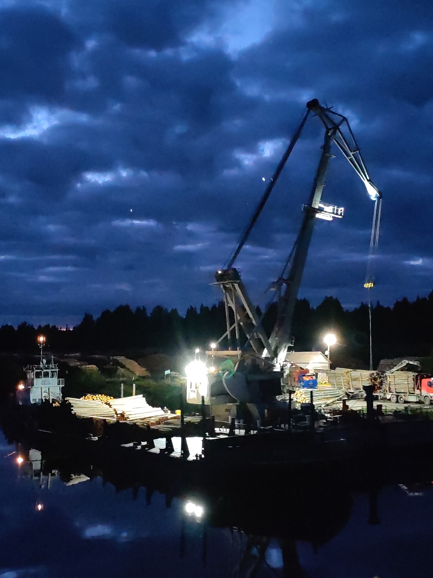 The image depicts a port scene in Annensky Most, Russia, at night. A large crane dominates the center of the frame, illuminated by several lights. The crane is positioned on a platform next to the water, with stacks of logs and lumber visible around it. A small tugboat is docked on the left side of the frame, and a truck is parked on the right. The sky is dark blue and cloudy, with a few visible stars. The water reflects the lights from the crane and surrounding structures, creating a mirrored effect.