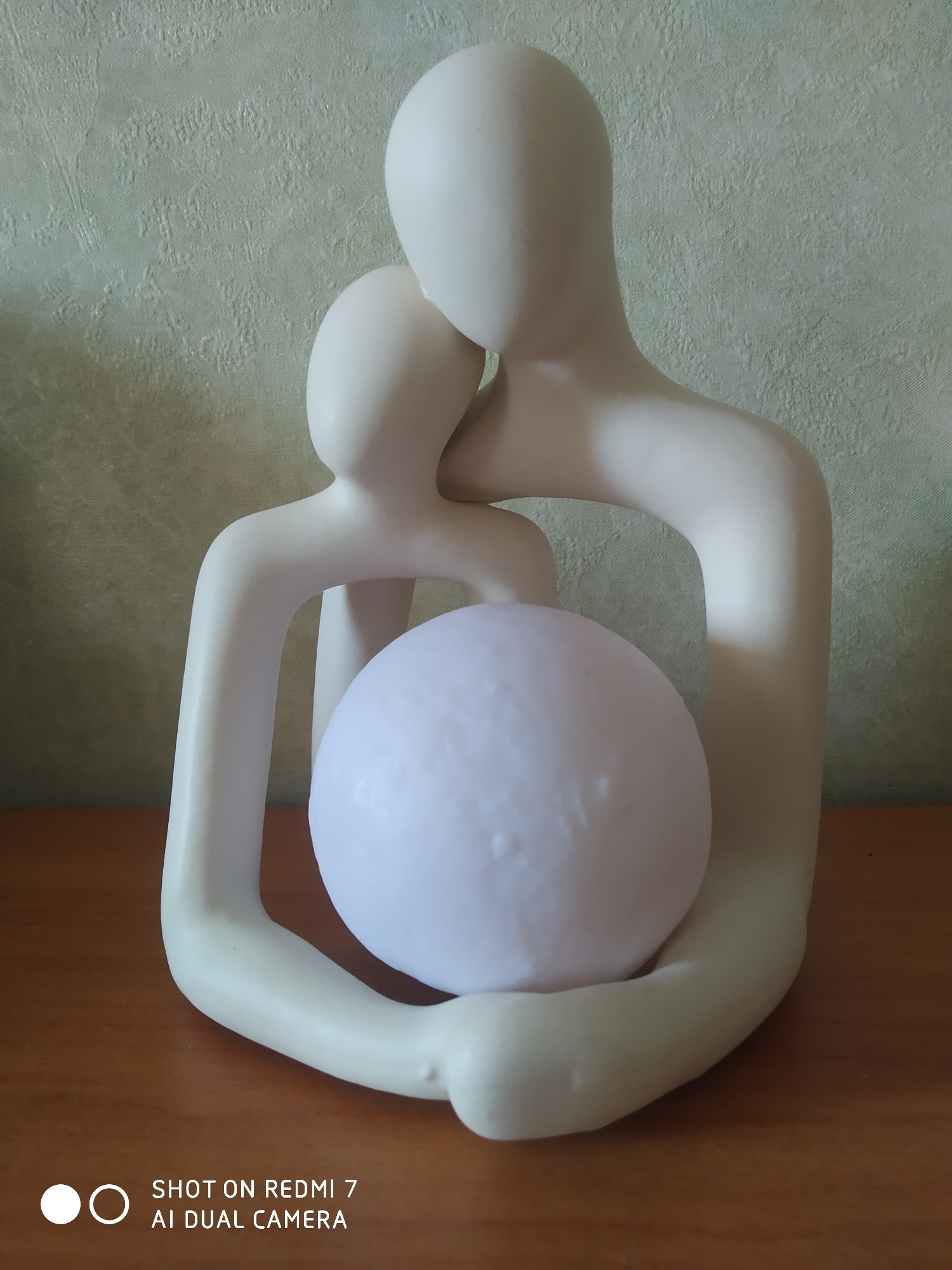 The image shows a white ceramic sculpture of two figures embracing. The figures are stylized and without detailed facial features. They are holding a spherical object in their arms. The sculpture is placed on a wooden surface. The background is a light green wall with a textured pattern. A watermark with the text "SHOT ON REDMI 7 AI DUAL CAMERA" is visible in the bottom corner of the image. The image was taken in the city of Chaikovsky, Russia.
