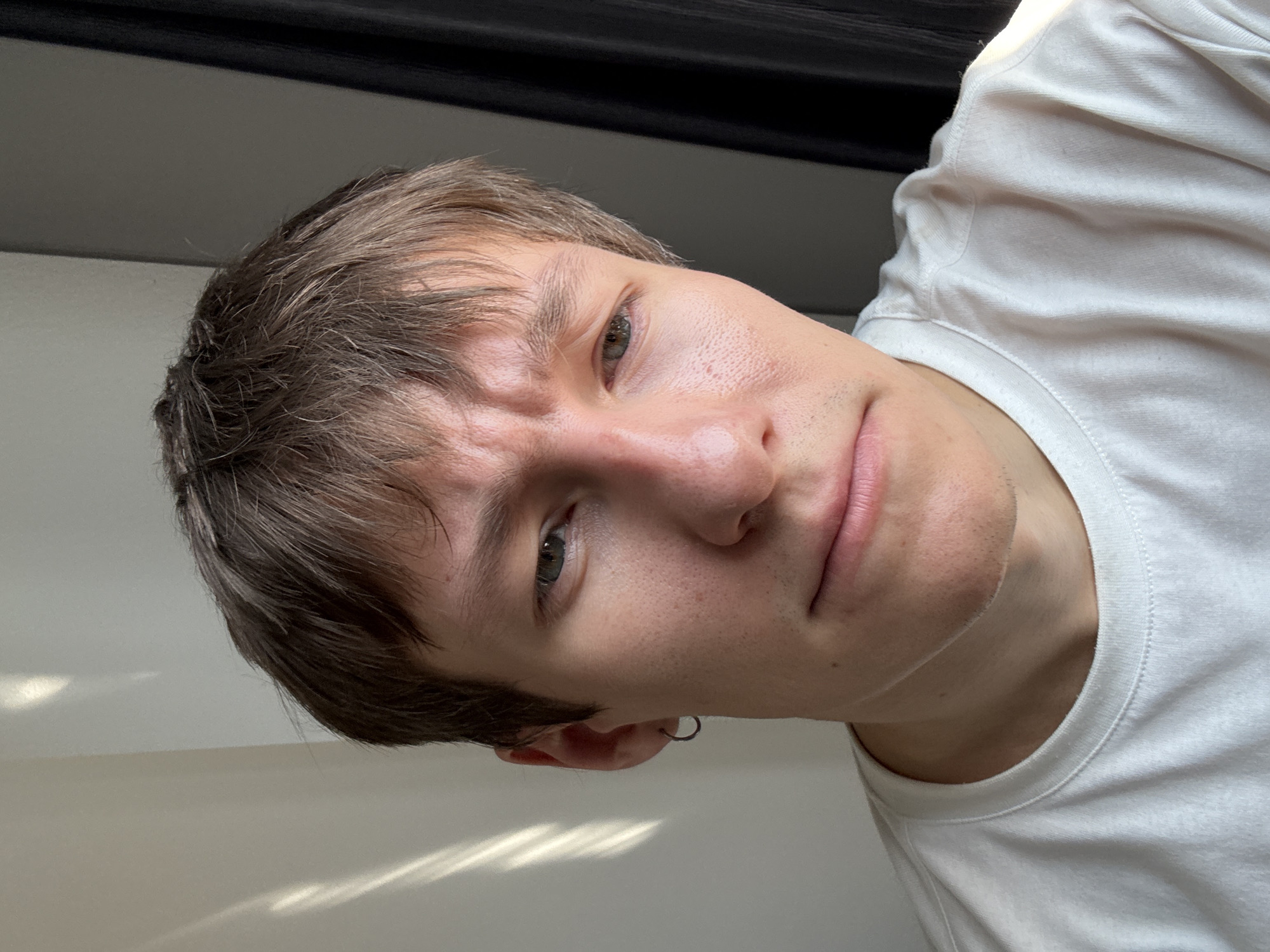A selfie of a fair-skinned young man with short brown hair, wearing a white t-shirt and a silver hoop earring. He is looking at the camera with a slight furrow in his brow and a neutral expression. The background consists of a light-colored wall and a dark, possibly black, area above. No evidence indicates this picture was taken in Hanoi, Vietnam.