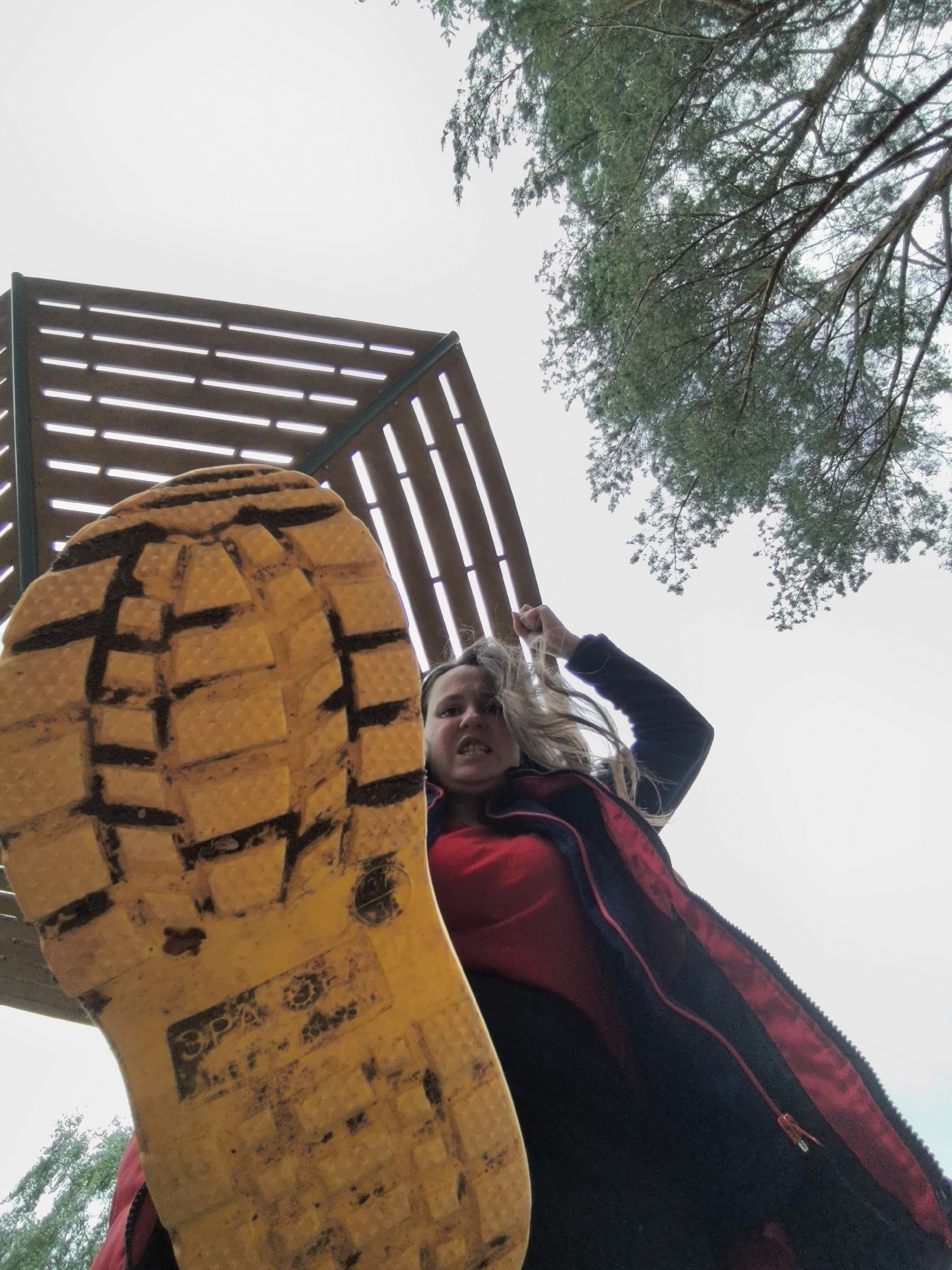 The image shows a low-angle view, looking up at a woman with blonde hair standing outdoors. A large, yellow boot sole with a tread pattern and dirt obscuring part of the view. The woman is wearing a black jacket with a red lining. A wooden structure with slats is visible in the background, along with green tree branches and a pale sky. The location appears to be a park or outdoor recreational area. The image location is Unknown, Unknown.