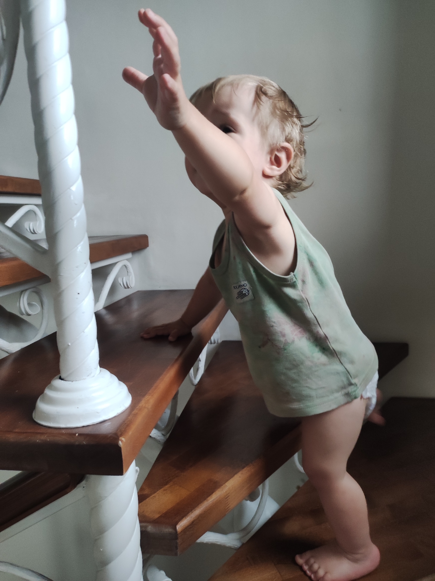 An indoor, eye-level, full shot depicts a young, fair-skinned child, approximately a toddler, in the act of ascending a wooden staircase. The child is positioned on the second step from the bottom, with the left hand resting flat on the dark brown wooden tread. The child's right arm is raised and extended upwards, with the hand open and fingers splayed, reaching towards an unseen point above and to the left of the frame. The child's head is turned slightly to the left, with the gaze directed upwards.
The child has light brown or blonde, slightly wavy hair and is wearing a light green sleeveless tank top with a faded, possibly tie-dye or mottled pattern, exhibiting creases. A white label with dark text and a graphic resembling a stylized hand is visible on the left side of the shirt, appearing mirrored. White underwear or a diaper is partially visible beneath the shirt. The child is barefoot, with the right foot resting on the second stair tread, while the left leg is bent, suggesting motion.
The staircase features dark brown wooden treads and white painted risers and decorative elements. A prominent white newel post or baluster with a spiral-fluted design stands vertically on the left, anchoring the lower part of the staircase. White scrollwork supports are visible beneath the wooden treads. The background consists of a plain, light grey wall. Lighting appears to originate from the right, casting mild shadows and highlighting the child's hair and raised arm. Minor imperfections are visible on the wooden treads and white painted surfaces of the stairs.