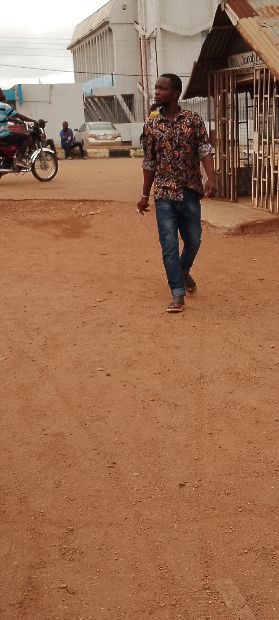 In Gboko, Nigeria, a man with dark skin and short hair walks on a dirt road. He is wearing a patterned short-sleeved shirt and blue jeans. His right hand is extended forward, holding a cigarette. The man is walking towards the right side of the frame. In the background, a white building with vertical lines is visible, along with a red motorcycle and a silver car. The environment appears to be a street in a town.