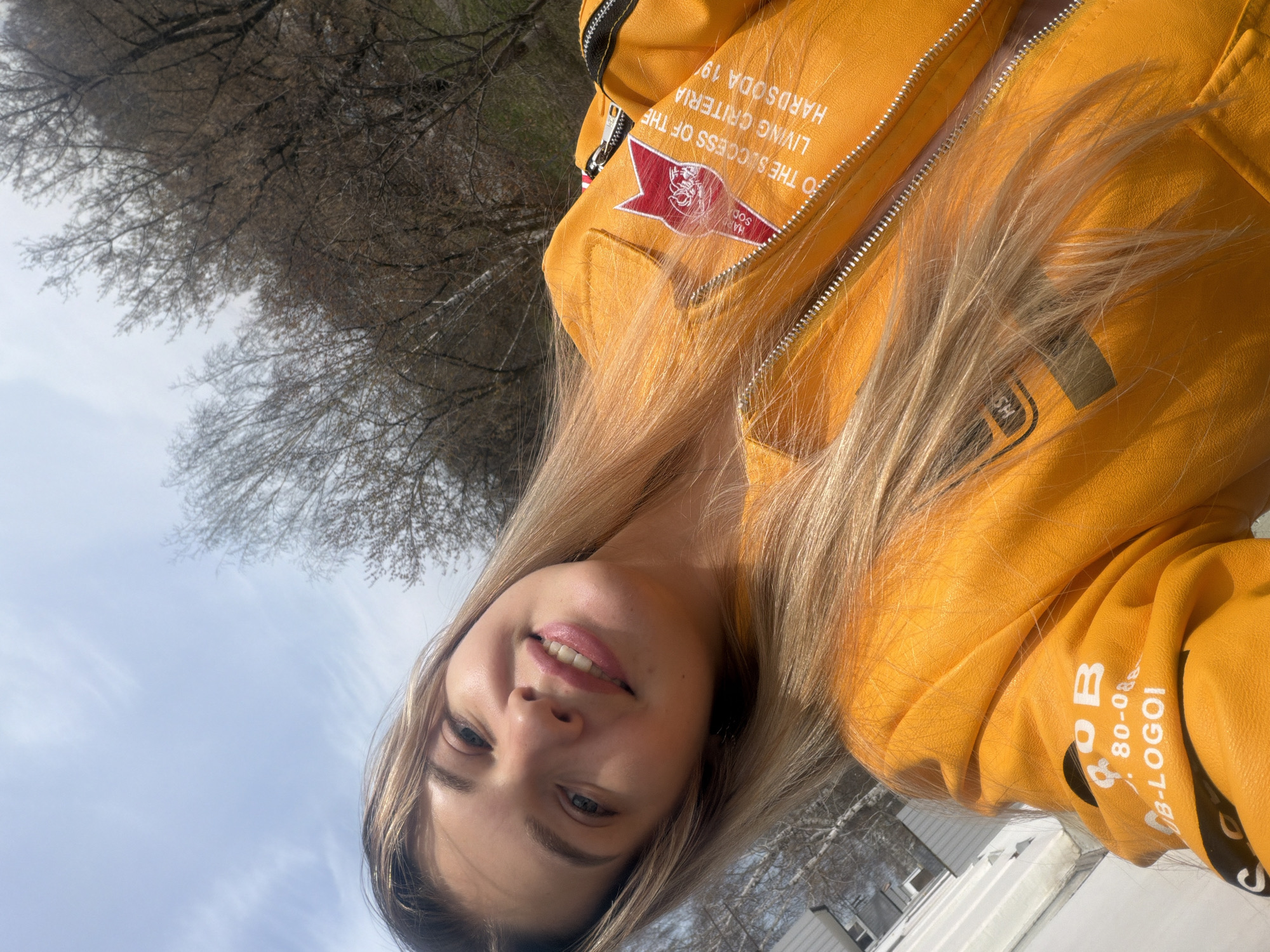 The image is a rotated selfie of a young woman with blonde hair wearing a yellow jacket. The jacket has text and a logo that includes the words "HARDSODA 19". The background consists of a blue sky, bare tree branches, and a portion of a light-colored building or structure. There is insufficient information to determine the exact location in Naberezhnye Chelny, Russia.