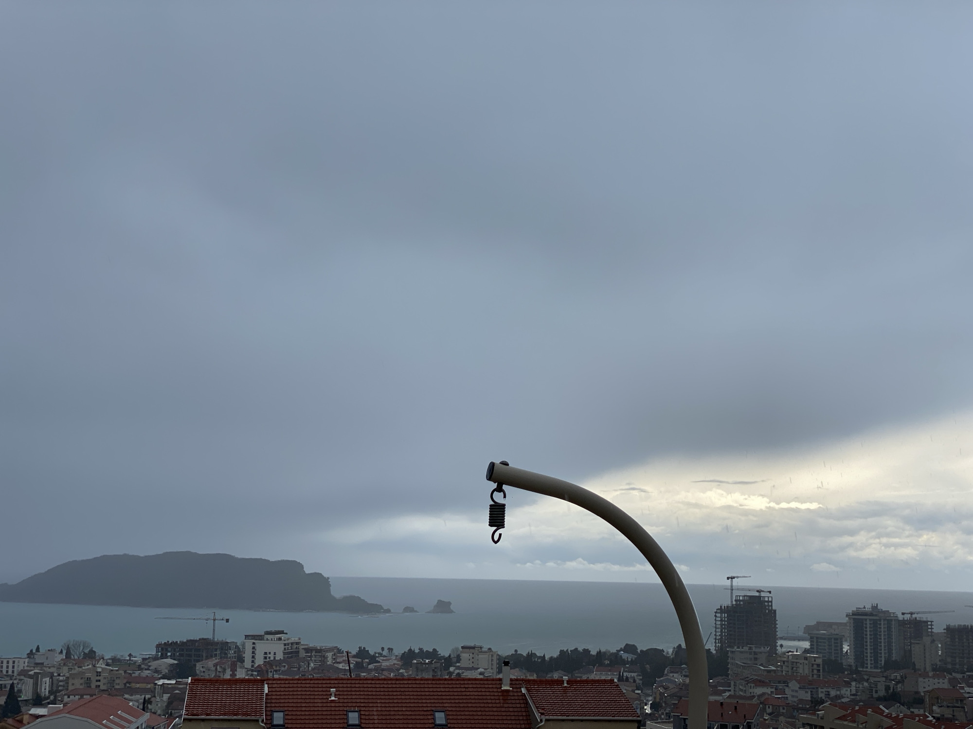 An elevated panoramic view captures the city of Budva, Montenegro, overlooking the Adriatic Sea under an overcast sky. In the immediate foreground, a light-colored, curved pole extends from the lower right, with a black metallic coil spring and hook suspended from its end. Below this fixture, red-tiled residential roofs are visible, some with skylight windows. The midground features a dense urban landscape of varied buildings, including several multi-story structures under construction, marked by exposed concrete frames and at least two tower cranes. Beyond the city, the teal-blue sea stretches to the horizon. On the left side of the background, a large, dark, mountainous island, identified as Sveti Nikola Island, dominates the seascape. Smaller rock formations are present in the water further right. The sky is predominantly grey and heavily clouded, with brighter patches visible towards the upper right, where faint streaks of precipitation are discernible. No individuals are depicted in the scene.