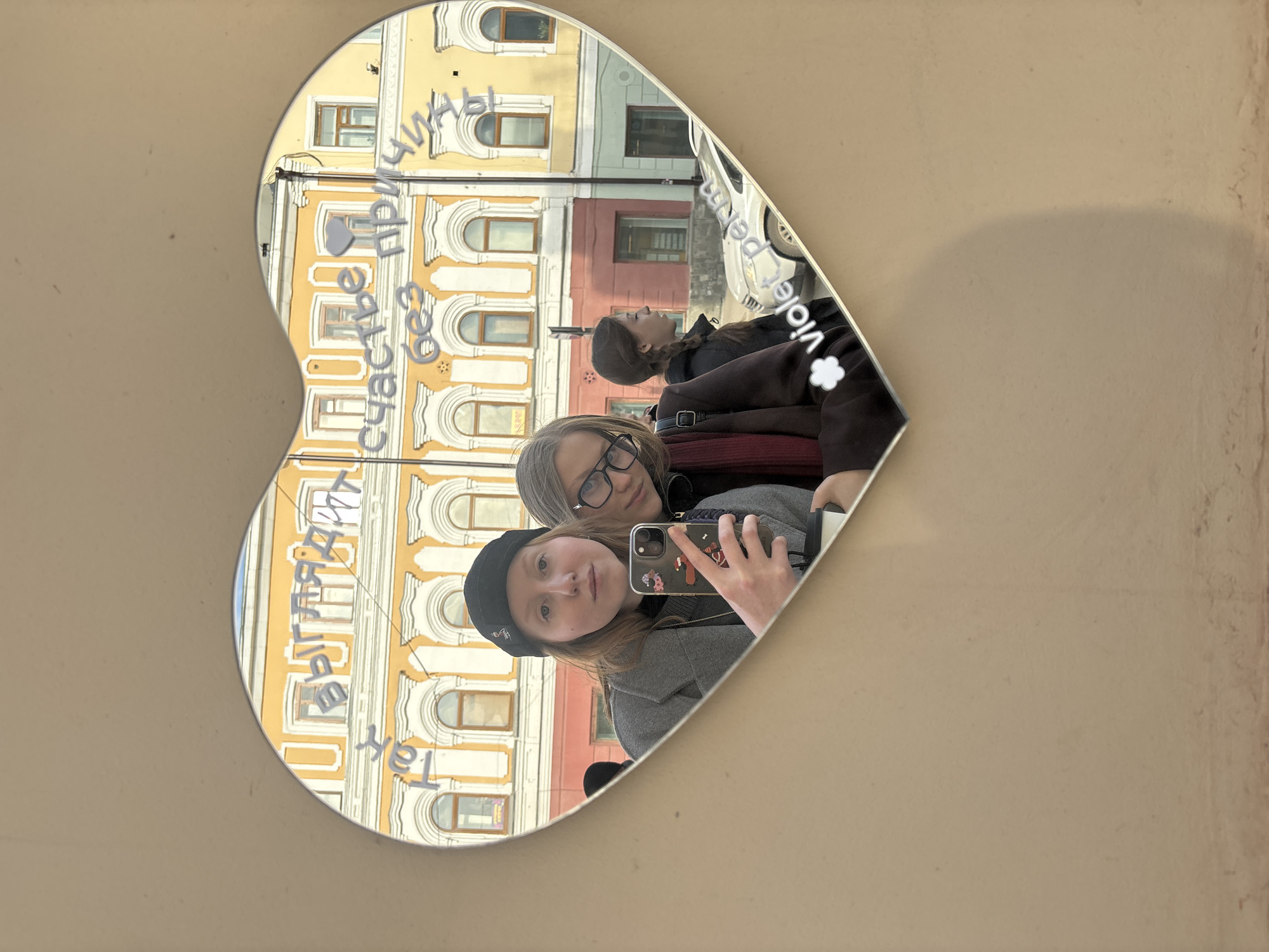 A heart-shaped mirror on a beige wall reflects three people and a building in Perm, Russia. The mirror shows a building with yellow facade and white arched windows. In the foreground, two people are visible: one wearing a black beanie and a grey jacket and the other with glasses and a red scarf. A third person with braided hair and a dark jacket is partially visible in the mirror behind them. The person in the beanie is holding a phone with a decorative case taking a selfie in the heart-shaped mirror. Writing is on the mirror, partially obscured by the reflection. A car is visible in the background reflected in the mirror.