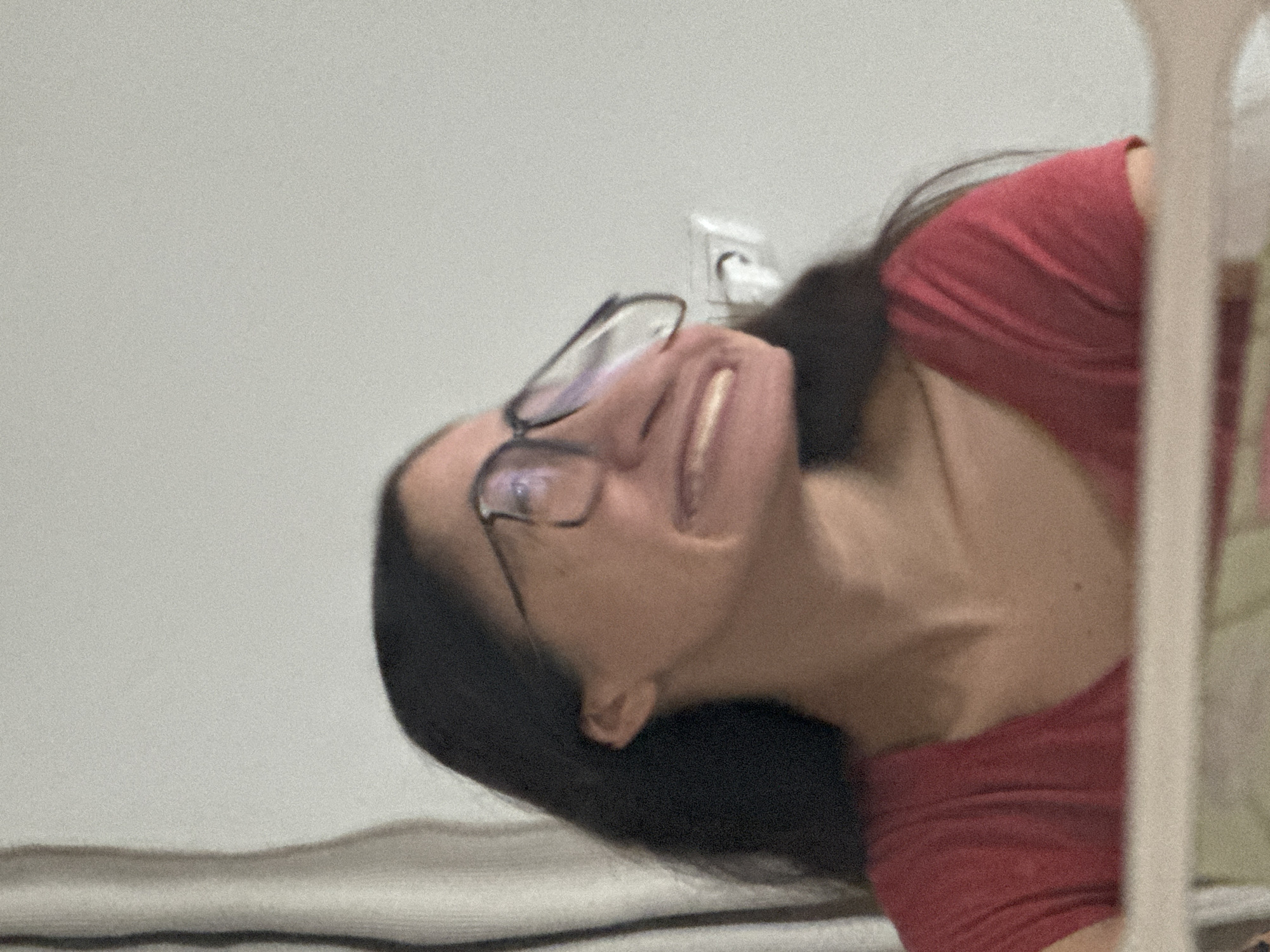 The image shows a woman with dark hair wearing glasses and a red shirt. She is looking up with a smile. Part of her body is obscured by a white, vertical object, potentially a door frame or part of furniture. In the background, there is a white wall with an electrical outlet. The image does not contain information to determine the location in Krasnoyarsk, Russia.