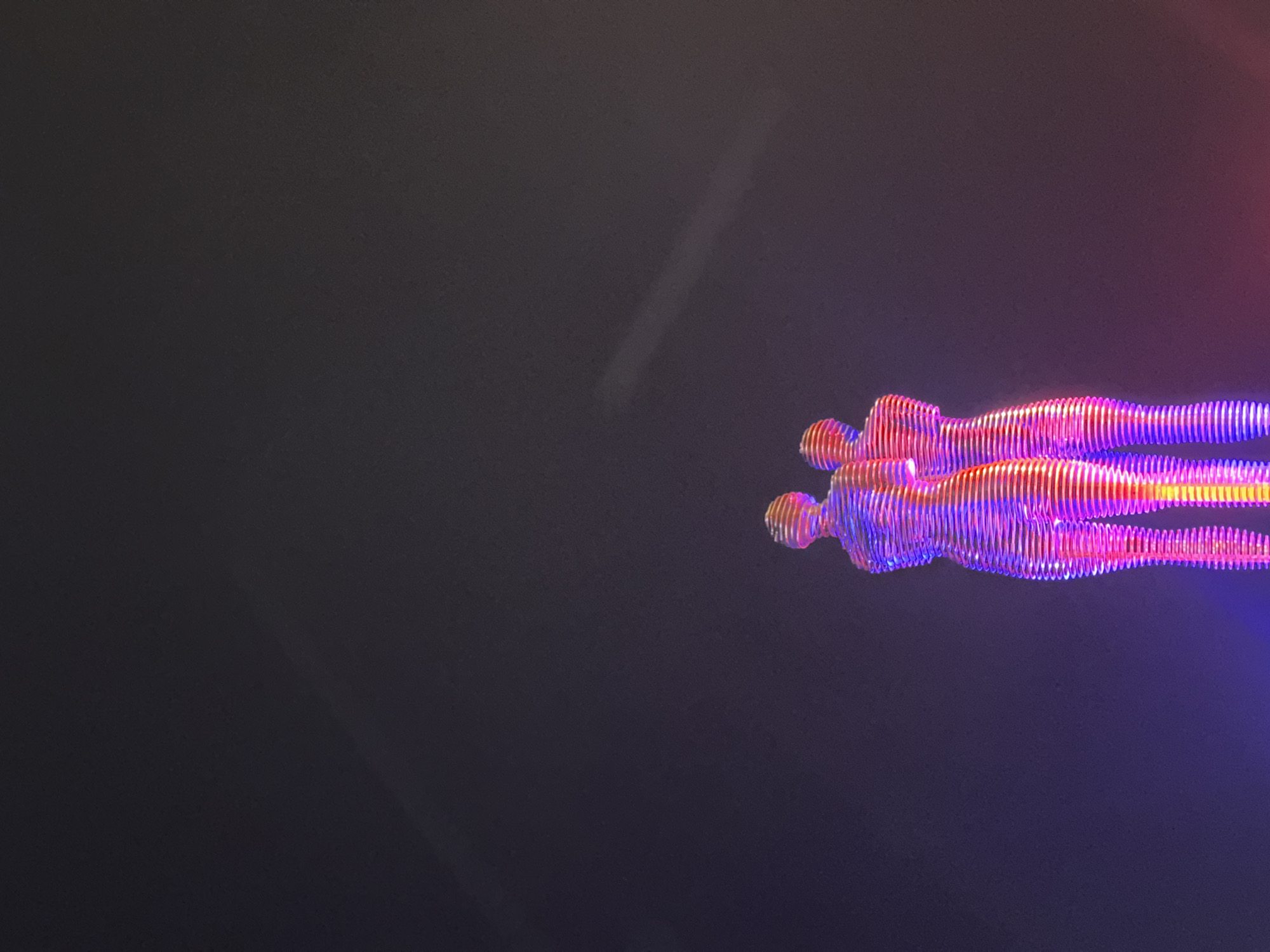 This image shows the Ali and Nino sculpture, a kinetic monument located on the shore of Batumi, Georgia, near the Black Sea. The sculpture is composed of two human figures, one male and one female, made of metal segments that are illuminated with colorful lights, primarily in shades of pink, purple, and blue. The figures are positioned facing each other, with their arms around each other in an embrace. They are designed to move, with the male figure gliding forward to embrace the female figure and then separating again. The sculpture is set against a dark, possibly night sky, with a faint, elongated light streak visible in the upper portion of the frame. The overall impression is one of romance and movement, highlighted by the vibrant lighting against the dark backdrop.