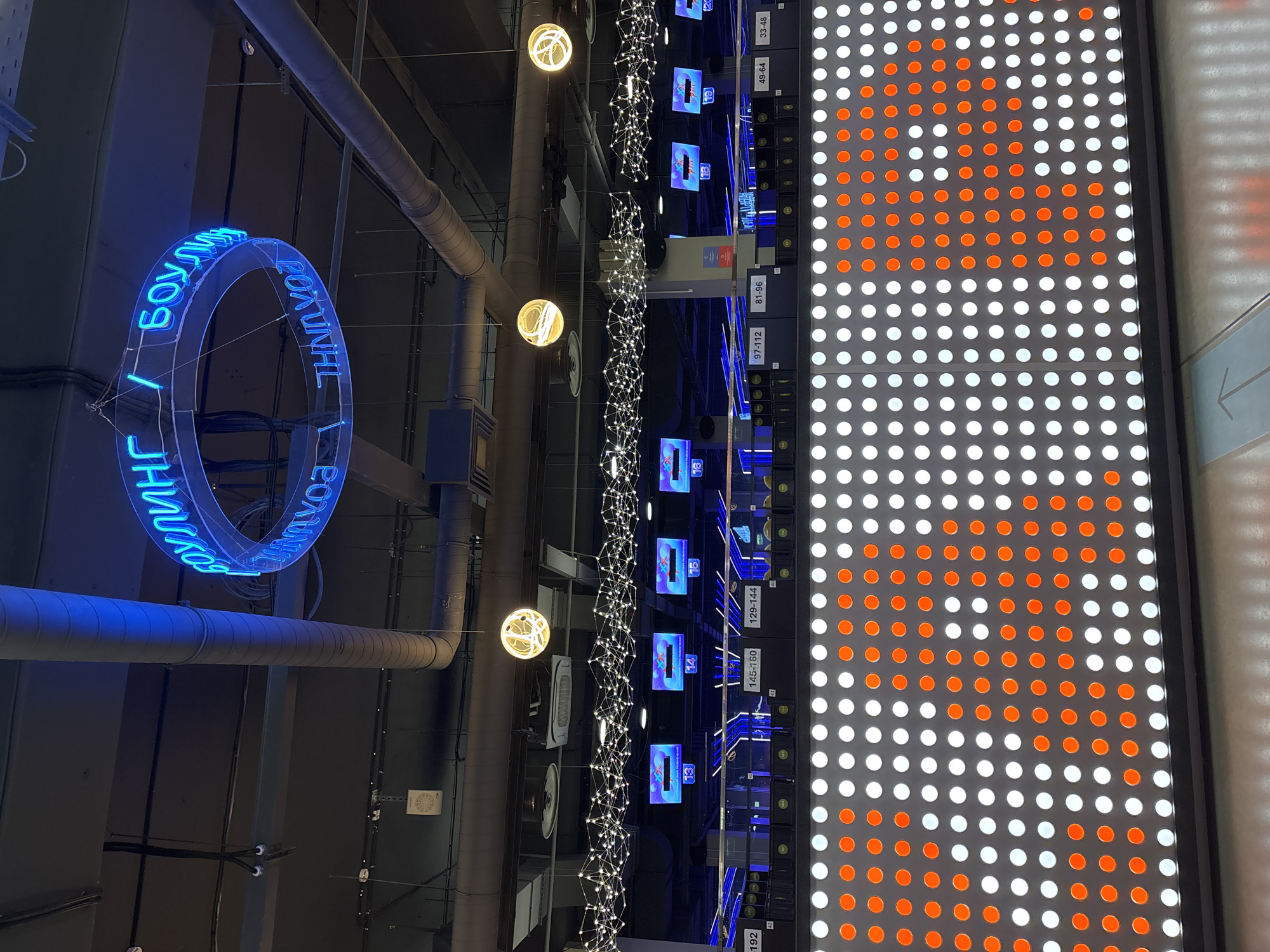 The image depicts the interior of a bowling alley in Saint Petersburg, Russia. A large, circular neon sign, glowing blue, hangs from the ceiling, displaying text that appears to be in Cyrillic script. Below the neon sign, a series of screens show lane numbers, such as "13", "14", "15", and "16". Further down, labels indicate ranges of lane numbers, for example, "145-160" and "129-144". The foreground is dominated by a wall covered in a grid of illuminated circles, alternating between white and orange, forming a large heart shape in the center. The environment is dimly lit, with industrial-looking pipes and ductwork visible on the ceiling, and strings of small white lights creating a decorative effect. No people are visible in the image.