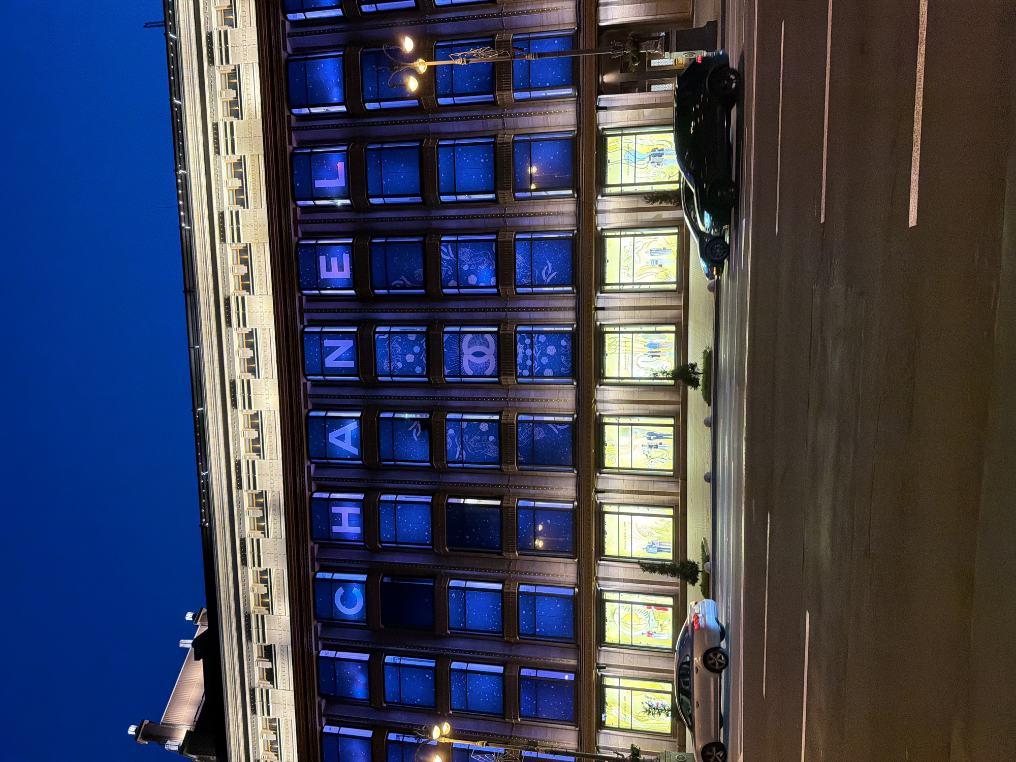 The image depicts the exterior of a building at night, illuminated by blue lights and internal yellow lighting. The building features large windows on multiple floors. The upper floor windows display the letters "C", "H", "A", "N", "E", "L" in large white lettering against a blue background with star-like patterns. One window also shows the Chanel logo. The lower floor windows are illuminated with yellow light and display mannequins dressed in clothing, suggesting it is a storefront.
In front of the building, a street is visible with several cars. On the left, a silver car is parked or moving slowly on the road. On the right, two dark-colored cars are also present on the road. The scene appears to be a commercial street in a city at night.
Based on the distinctive architectural style and the Chanel store facade, this location is identified as the Chanel store on Khreshchatyk Street in Kyiv, Ukraine. Khreshchatyk Street is a major thoroughfare in Kyiv, known for its architecture, shops, and public spaces. The building itself is a historical structure, often referred to as the "House with Chimeras" or its neighboring buildings on Khreshchatyk are significant landmarks. The presence of cars and the illuminated street further confirm an urban setting.