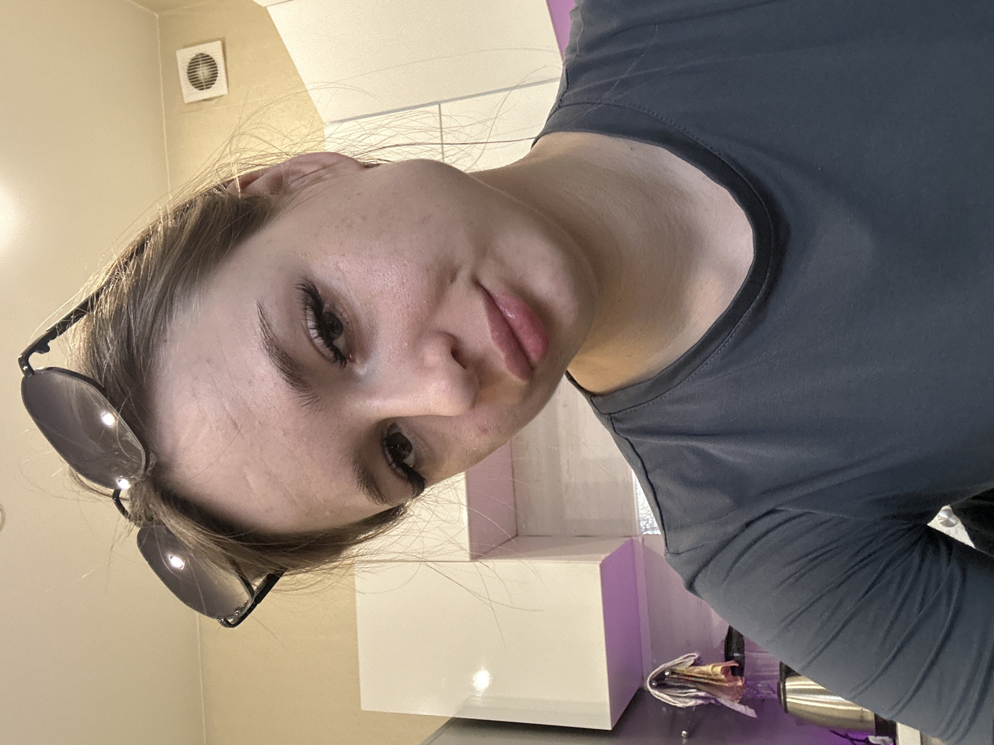 The image shows a close-up selfie of a fair-skinned person with light brown hair. The person is wearing a gray t-shirt and has sunglasses perched on their head. The background appears to be a modern interior, possibly a kitchen or bathroom, with white and light purple cabinets or shelving. A silver object is visible on a counter, likely a coffee machine or similar appliance. A vent is visible on the upper-left wall. The exact location and city are unknown.