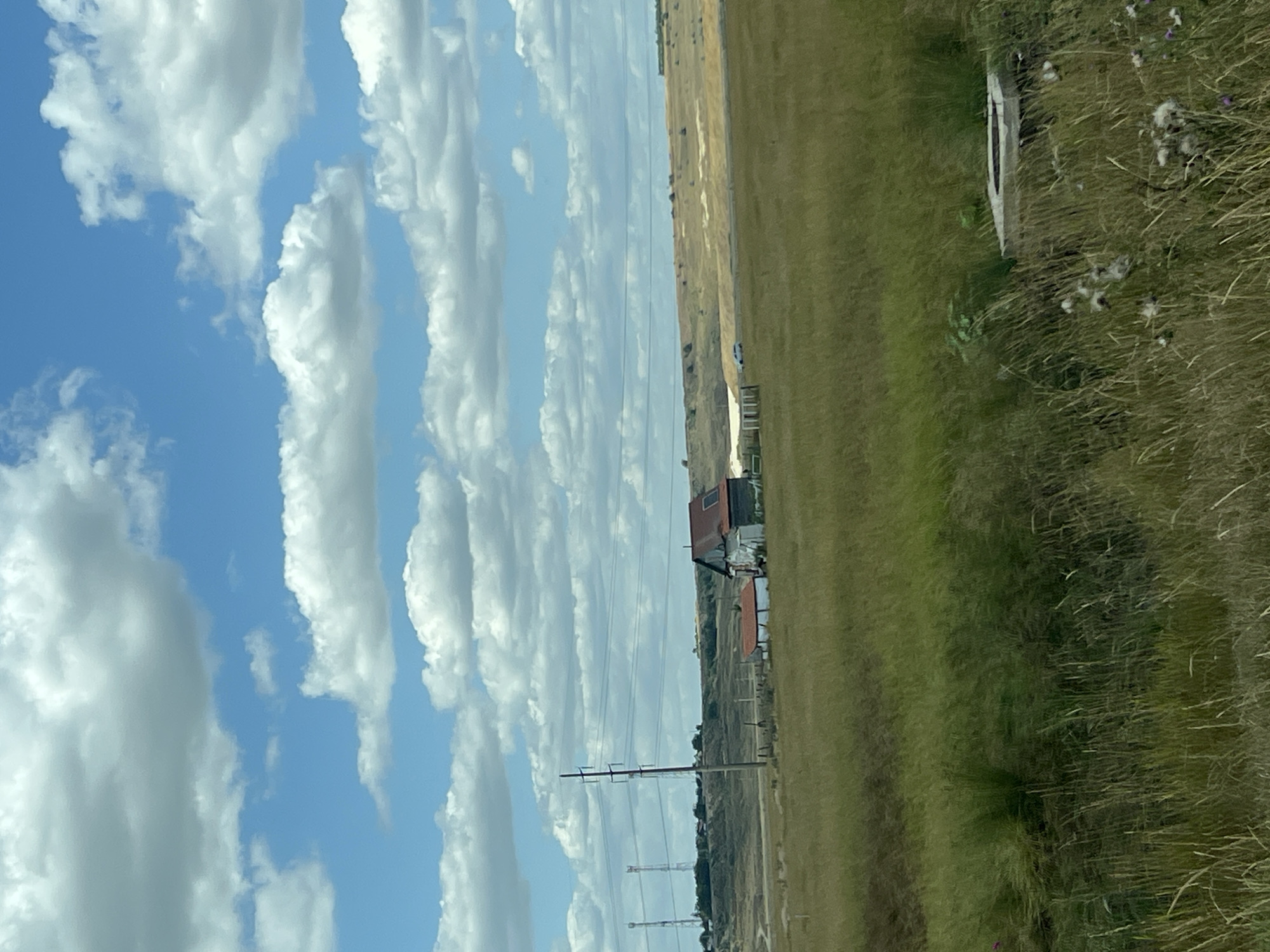 The image depicts a rural landscape under a cloudy blue sky. In the mid-ground, a cluster of buildings, possibly a farmstead, is situated amidst a field of dry grass. One prominent structure has a red roof and appears to be weathered. To the left of the buildings, utility poles with visible wires stretch across the scene. In the background, rolling hills are visible, with some areas appearing parched or eroded. The foreground is dominated by tall, dry grass and some wild vegetation. A concrete structure, possibly a well cap or a base for equipment, is visible in the lower right corner. The overall impression is of an arid, open rural environment. The location is identified as Podu Iloaiei, Romania, suggesting a setting in the Romanian countryside.