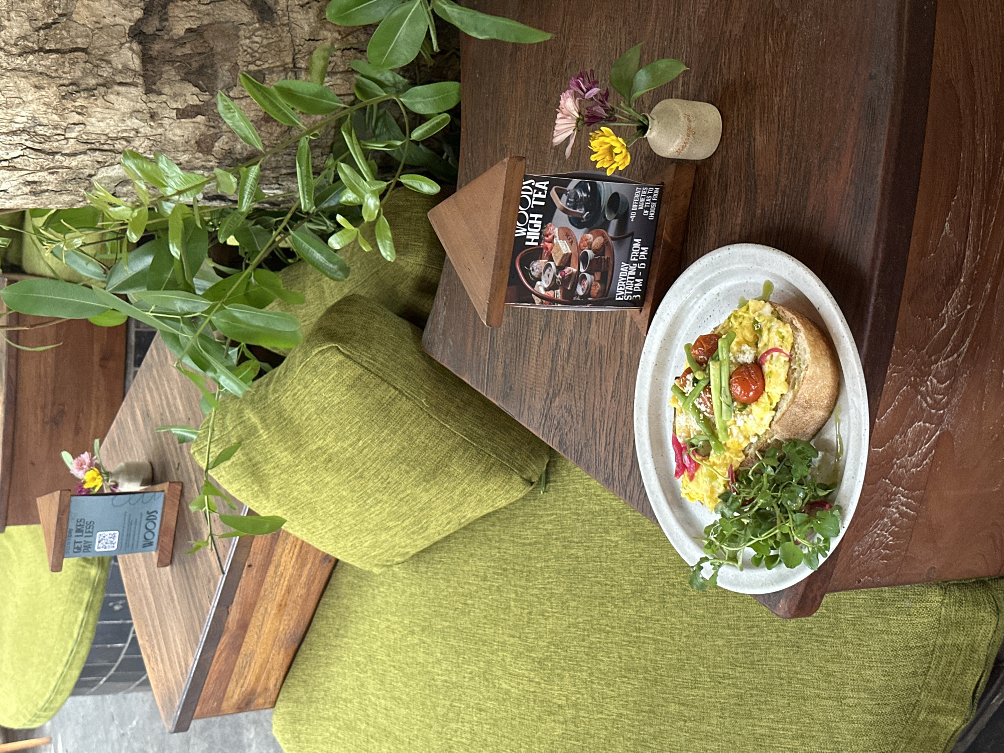 This image captures a serene cafe setting, likely in Mengwi, Indonesia, with a focus on a meal served on a wooden table. The scene is shot from a top-down perspective, slightly angled.
The main subject is a plate of food, featuring what appears to be scrambled eggs on a slice of bread, garnished with halved cherry tomatoes, green asparagus or beans, sliced radishes, and a generous portion of fresh greens like watercress. A drizzle of oil adds to the presentation. The plate is white with a textured rim.
Beside the plate, on the dark wooden table, is a wooden display card advertising "WOODS HIGH TEA." The card details that the high tea is offered "EVERYDAY STARTING FROM 3 PM - 6 PM" and includes "+40 DIFFERENT VARIETIES OF TEAS TO CHOOSE FROM." It also features an image of a high tea spread. A small, light-colored vase holding a few colorful flowers (yellow and purple) adds a touch of natural beauty to the table. Another smaller wooden display card is visible further away, displaying "GET LIKES PAY LESS" and "WOODS" along with a QR code, suggesting a promotion or social media interaction.
The table is surrounded by comfortable-looking green upholstered seating, contributing to a relaxed atmosphere. Lush green plants, possibly from a tree trunk integrated into the decor, are prominent on the left side of the frame, enhancing the natural and tranquil ambiance of the cafe. The combination of natural wood, vibrant greenery, and soft seating creates an inviting environment for enjoying a meal or afternoon tea.