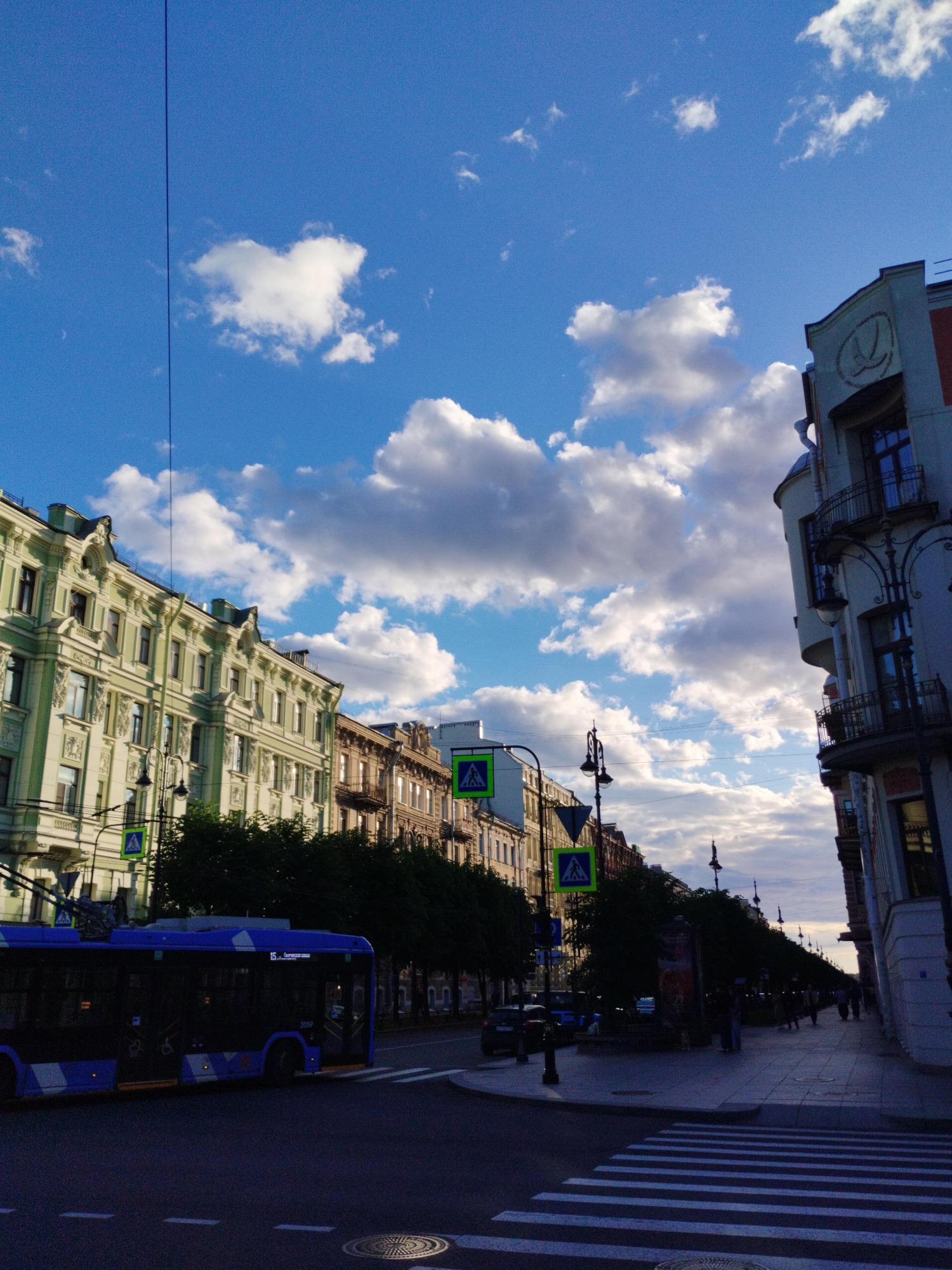 The image depicts a street scene in Saint Petersburg, Russia, likely in the historic center. A blue trolleybus, displaying the number "15" and destination "Сенная пл.", is captured mid-transit on the left side of the frame. In the background, a row of ornate, historic buildings with multiple stories lines the street, showcasing architectural details like balconies and detailed facades. Green trees are planted along the sidewalk in front of these buildings, creating a verdant canopy. Pedestrian crossings with white stripes are visible on the asphalt road, and streetlights with elaborate designs punctuate the sidewalk. The sky is a vibrant blue, adorned with fluffy white clouds, suggesting fair weather. A few individuals are seen walking on the sidewalk on the right side of the image, appearing as silhouettes against the slightly darker backdrop of the buildings and pavement. The overall impression is of a typical urban streetscape in Saint Petersburg, combining historical architecture with active city life.