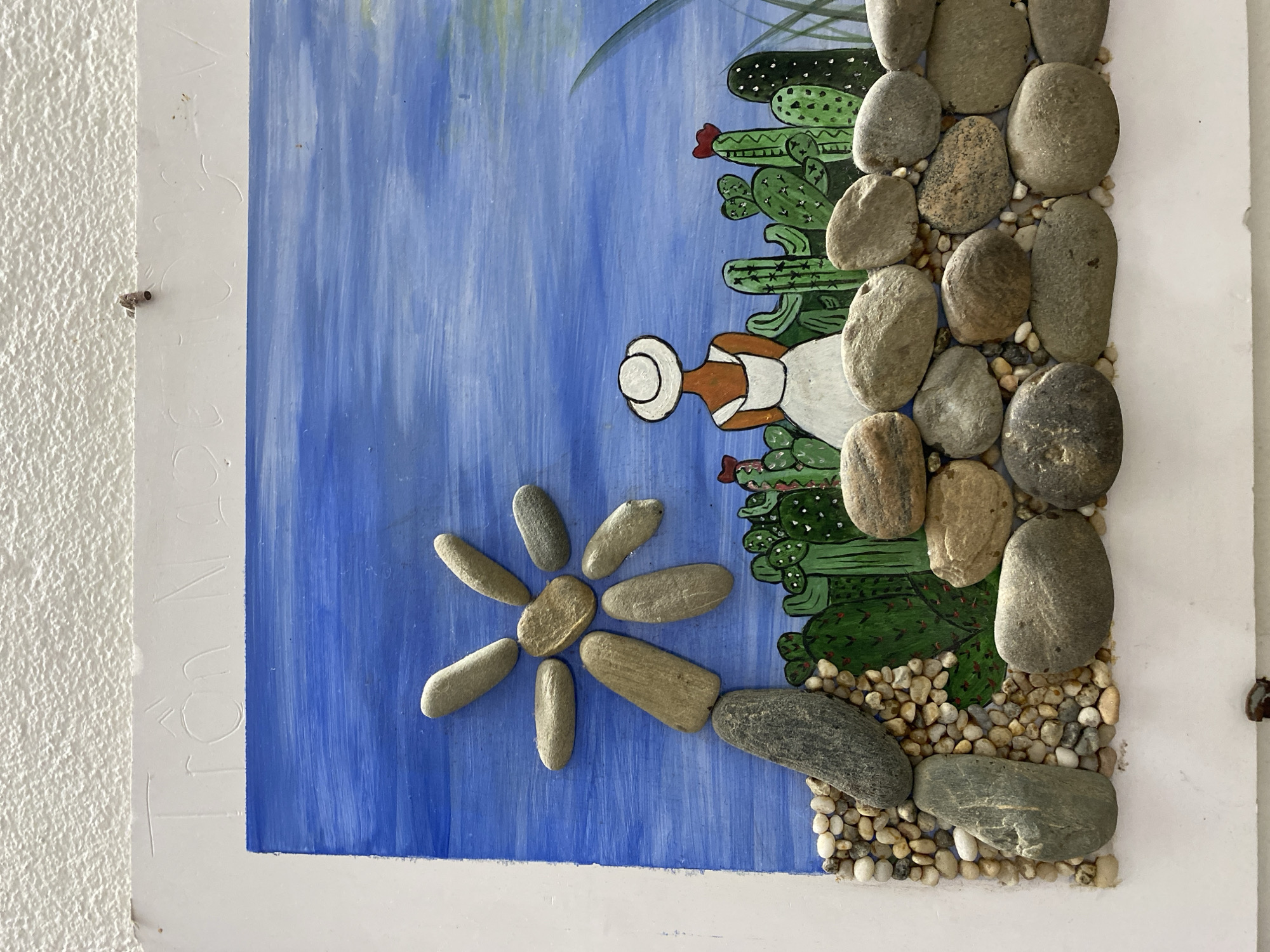 This is a close-up, eye-level shot of a mixed-media artwork mounted on a white surface, likely a wall. The artwork depicts a desert scene with a bright blue sky. In the foreground, a stone mosaic forms a rough wall of various sized pebbles and larger river stones. Above this wall, painted green cacti with red flowers are depicted. To the left, a collection of smooth, oval stones are arranged to resemble a sun or flower. In the center-right of the scene, a person viewed from the back, wearing a white hat and a white dress, stands amongst the cacti. The artwork is signed at the top with scratched-in letters that appear to read "Trân Ngoc Quý". A small, dark object, possibly an insect cocoon or a piece of debris, is attached to the white surface above the artwork. The texture of the white surface appears to be rough and stucco-like, suggesting it is a wall. The artwork is likely located in Đà Nẵng, Vietnam, given the Vietnamese name signed on it.
