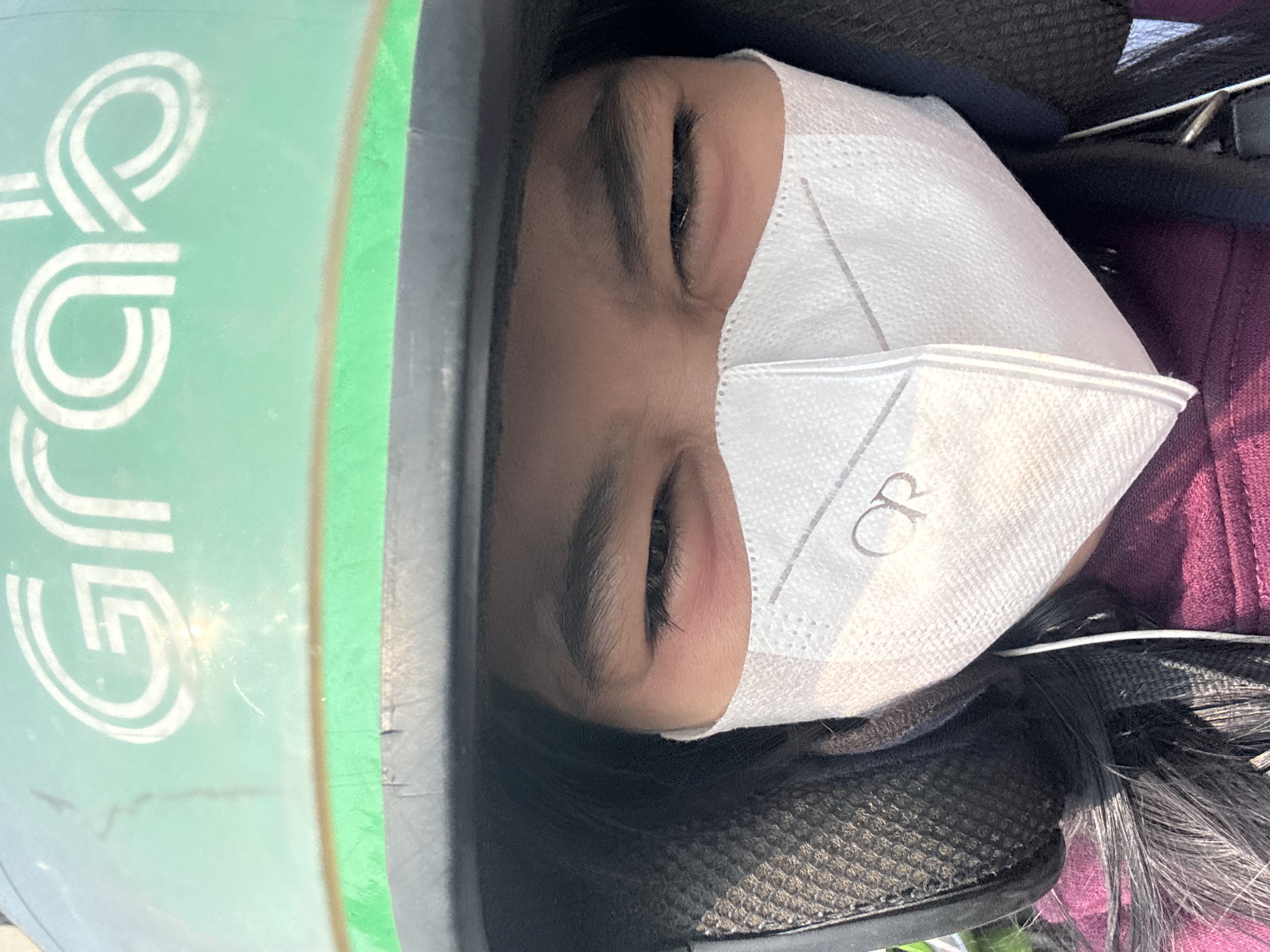 A person wearing a green Grab helmet and a white mask is looking at the camera. The person's eyes are narrowed, and they appear to be in sunlight. The helmet has the Grab logo on it. The person is wearing a maroon shirt and appears to be on a motorcycle. The location is North Jakarta, Indonesia.