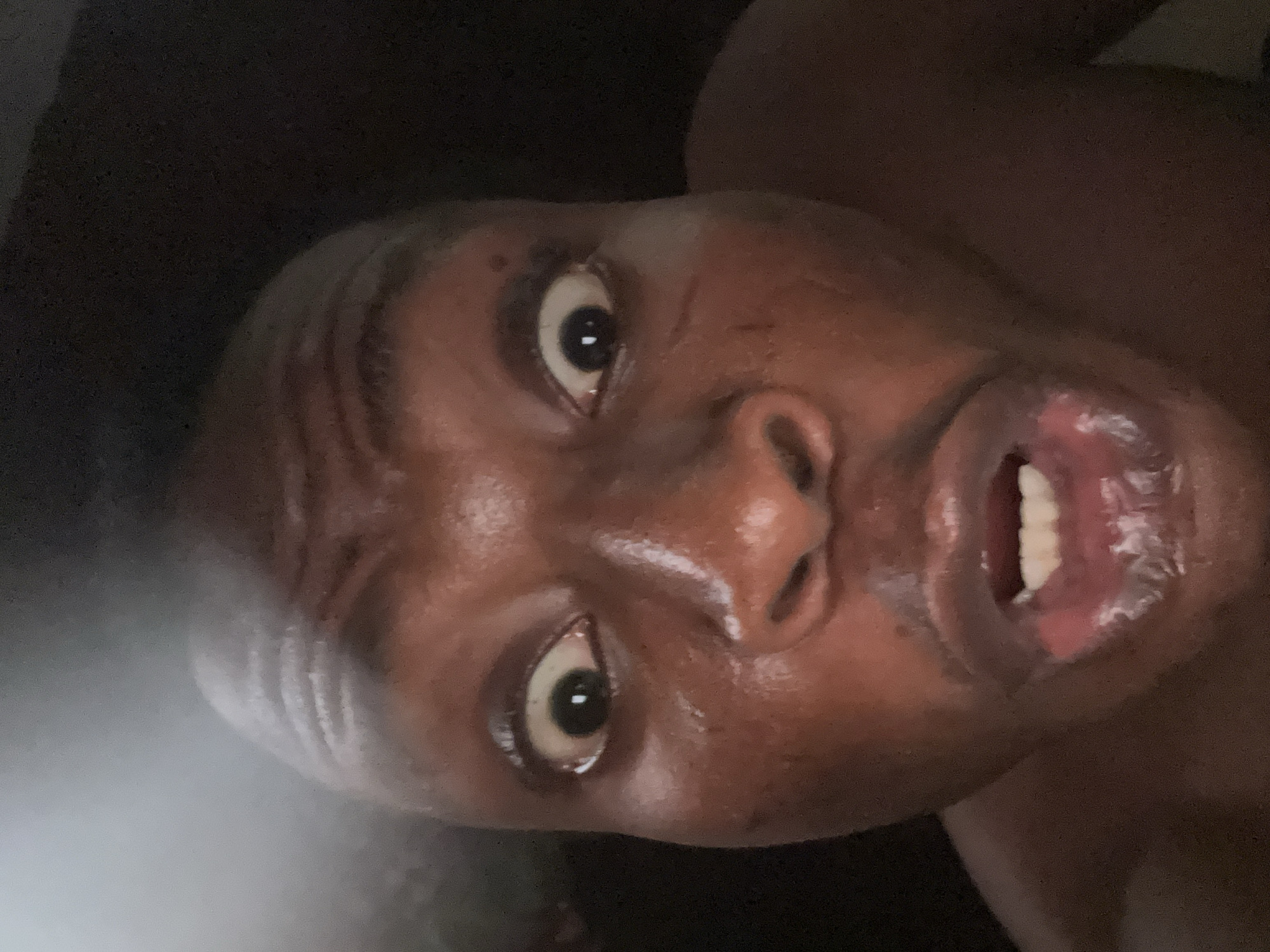 A close-up, tilted view shows a dark-skinned person with wide eyes and an open mouth. The person's face appears sweaty and shiny. The background is dark and indistinct. The location is unknown.