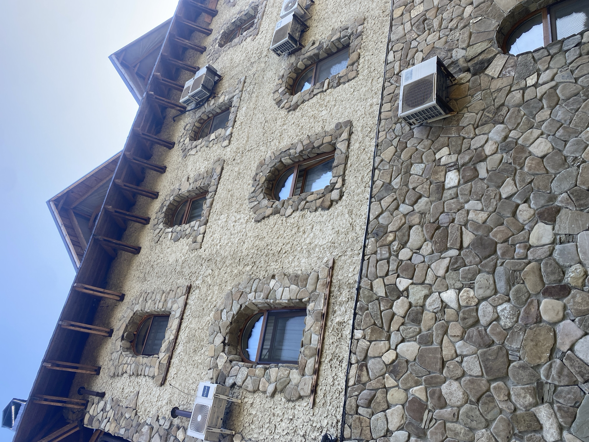 The image displays the exterior of a multi-story building constructed with rough-hewn stone at the lower levels and a textured, light-colored stucco on the upper floors. The architecture features arched windows with stone surrounds and wooden eaves with exposed rafters. Several air conditioning units are mounted on the exterior walls. The scene is an outdoor view looking upwards towards the building against a clear blue sky. The location is identified as Goryachiy Klyuch, Russia, specifically the building that houses the Goryachiy Klyuch Museum of Local Lore.