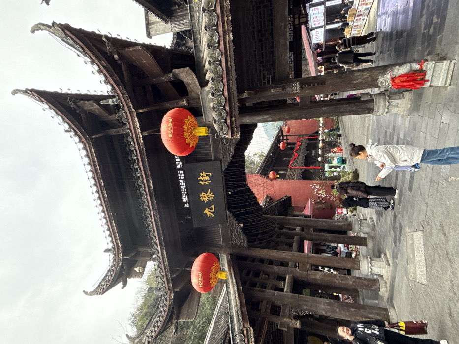 The image displays an outdoor scene at Jiuli Street in Pengshui Miao and Tujia Autonomous County, China. The foreground is dominated by a large, dark-stained traditional Chinese archway, characterized by multiple tiers of curved tiled roofs and numerous substantial wooden support pillars resting on stone bases. A black rectangular sign with golden Chinese characters "九黎街" (Jiulijie) is centrally suspended from the archway. Two round red lanterns with golden patterns and yellow tassels hang from the archway, one on each side of the sign. Small string lights are visible hanging beneath the archway structure.
Beyond the archway, a paved pedestrian street extends into an area lined with traditional multi-story buildings, some featuring prominent red-painted walls and dark roofs. These buildings are decorated with additional red lanterns and red fabric streamers. Green, tree-covered hills or mountains are visible in the background.
On the left side of the foreground, a woman in a black jacket with white lettering and dark pants holds a yellow and black striped shopping bag, her mouth slightly open. To the right, another woman, dressed in a light-colored jacket and blue jeans, with her hair in a bun, faces right and extends her right arm, pointing forward. In the mid-ground, under the archway's left side, a group of three individuals appears to be conversing. Further down the street on the right, additional people are visible, including one man in a striped shirt. The sky is overcast. A small stone lion statue with red ribbons is present at the base of a right pillar.