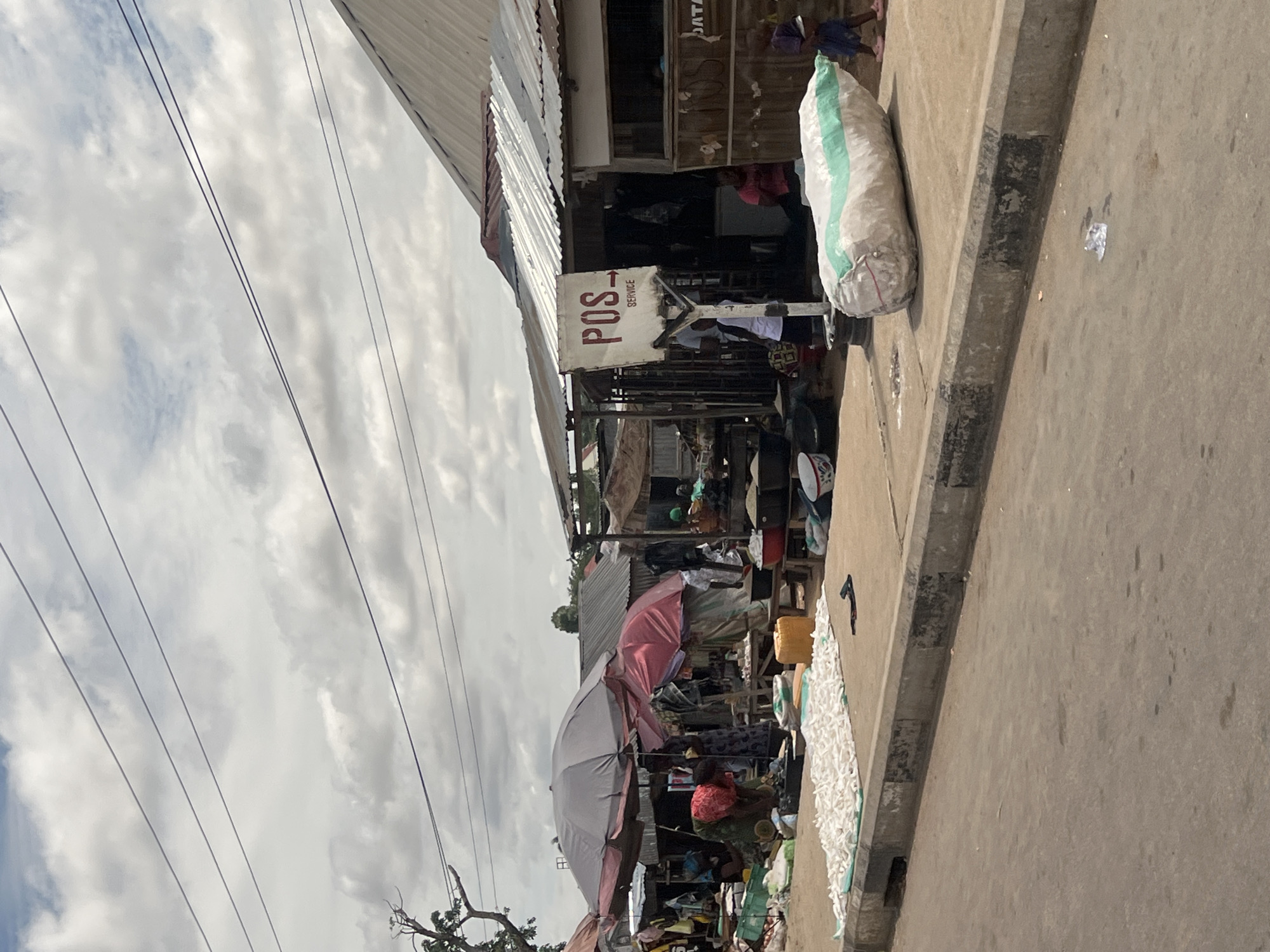 This image depicts a street market scene in Jalingo, Nigeria, under an overcast sky with visible power lines traversing the upper frame. The foreground features an asphalt road with scattered debris and a concrete curb separating it from a paved sidewalk. On the sidewalk, a large, elongated white sack with a green stripe rests, along with a mat displaying numerous small, white, elongated objects, possibly dried fish or food items. A dark object, possibly a sandal, lies near the mat.
The midground is dominated by a series of informal market stalls and structures with corrugated metal roofs. Two large open umbrellas, one grey and one red, provide shade over vendor areas. Within these stalls, several individuals are visible: one person in a red headwrap is bent over a display, interacting with items. Other people are standing or moving between stalls. A prominent white sign, supported by a metal post, reads "POS SERVICE" in red lettering with an arrow pointing right. Near this sign, an adult in a white shirt is partially visible, and a child in a blue garment is walking past the large white sack. Various market goods, including bowls containing grains or food, are arranged on low wooden tables and directly on the ground.
The background shows additional rudimentary structures and sparse tree branches against the cloudy sky. The overall scene suggests an active outdoor commercial area.