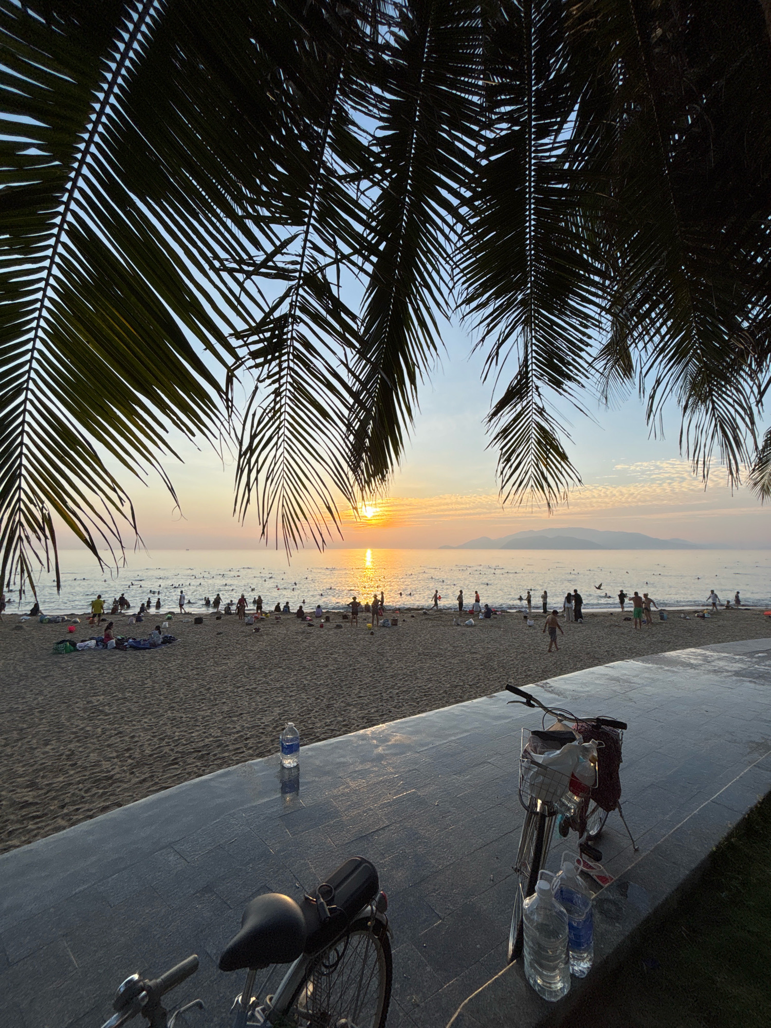 This image depicts a beach scene in Nha Trang, Vietnam, likely during sunset or sunrise. The foreground features a paved walkway with two bicycles parked on it. One bicycle is closer to the viewer and is equipped with a basket filled with items, and two large bottles of water are placed at its base. The other bicycle is partially visible behind it. Beyond the walkway, a sandy beach is visible with numerous people engaged in various activities, including swimming in the ocean and relaxing on the sand. Palm fronds hang into the frame from the top, suggesting the location is near a coastal area with palm trees. The ocean stretches to the horizon, where a landmass is faintly visible. The sky displays soft hues of orange and yellow, indicative of a sunset or sunrise, with some scattered clouds. The overall atmosphere is serene and recreational.