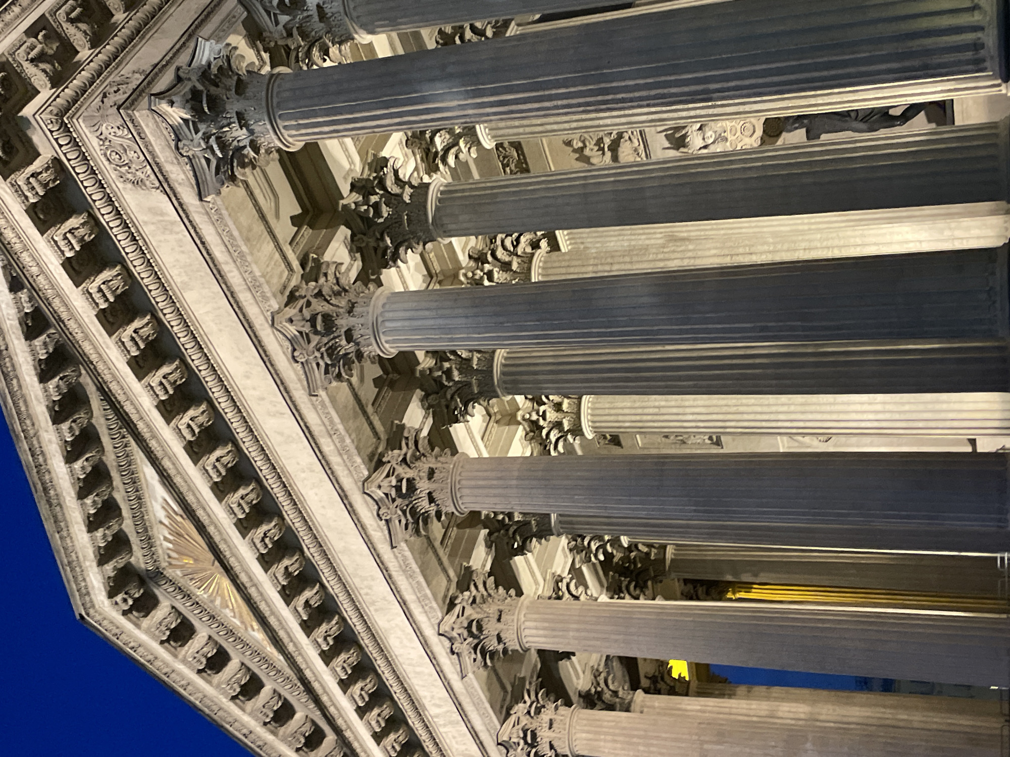 The image captures the facade of a grand neoclassical building in Saint Petersburg, Russia, at dusk or night. The sky above is a deep twilight blue. The primary focus is on the monumental Corinthian columns that support a classical pediment adorned with intricate carvings and a sunburst motif. The columns are illuminated from below, casting dramatic shadows and highlighting their fluted surfaces and ornate capitals. Between some of the columns, glimpses of interior architectural details and possibly a statue are visible, suggesting a large public building. The overall impression is one of grandeur, historical significance, and architectural beauty.