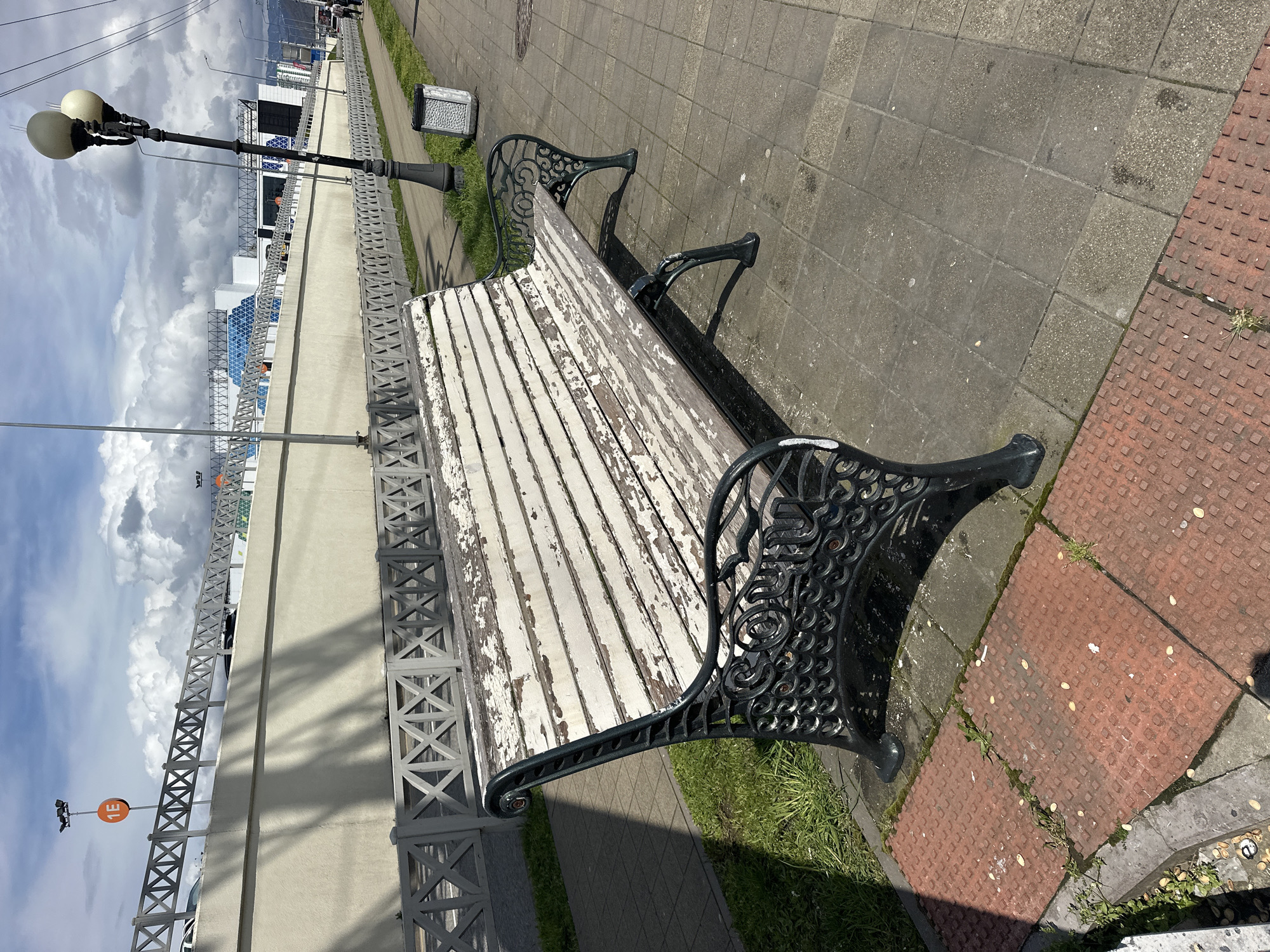 A high-angle, outdoor daytime shot shows a weathered, white-slatted bench with black ornate metal supports on a paved surface, likely a sidewalk or promenade. The bench, with peeling paint, sits next to a raised concrete barrier with a decorative metal fence. Beyond the barrier, a modern architectural structure with blue glass accents is visible against a cloudy sky. Street lamps are positioned along the barrier. The pavers surrounding the bench are a mix of gray concrete and red textured slabs. The scene is likely a public space in Sochi, Russia, based on the architecture and infrastructure. The image contains no people.