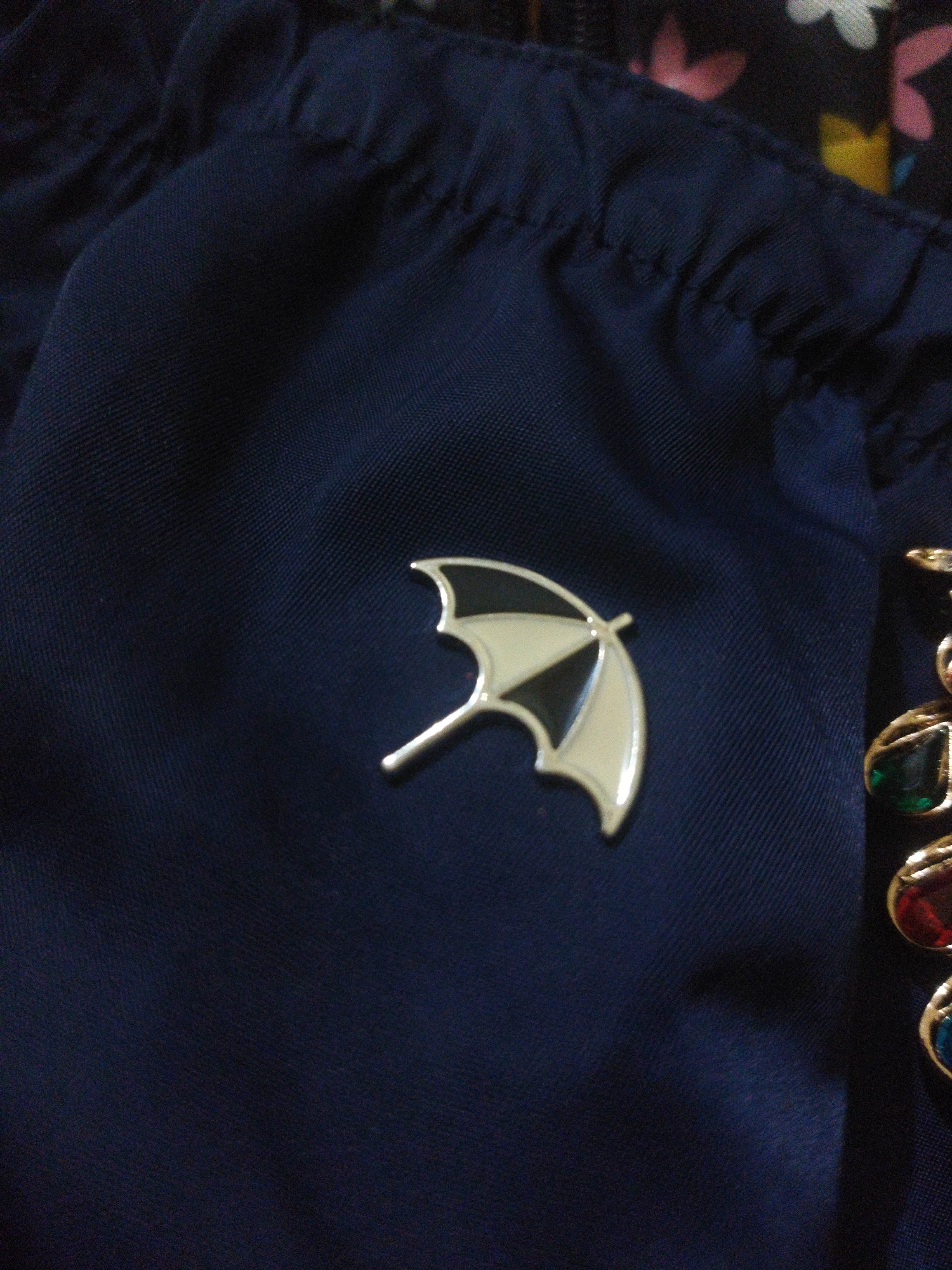A close-up, top-down shot shows a navy blue fabric with a textured weave, likely part of a garment or bag. A small, silver and black enamel pin shaped like an open umbrella is affixed to the fabric. To the right of the umbrella pin, partially visible, is a gold-colored jewelry item with colorful gemstones, including green, red, and blue, set in a decorative pattern. The background is out of focus, showing hints of a floral pattern with pink, white, and yellow elements. The lighting suggests an indoor environment, possibly a home or retail setting. There is no discernible information about the city of Suci, Indonesia, in this image.