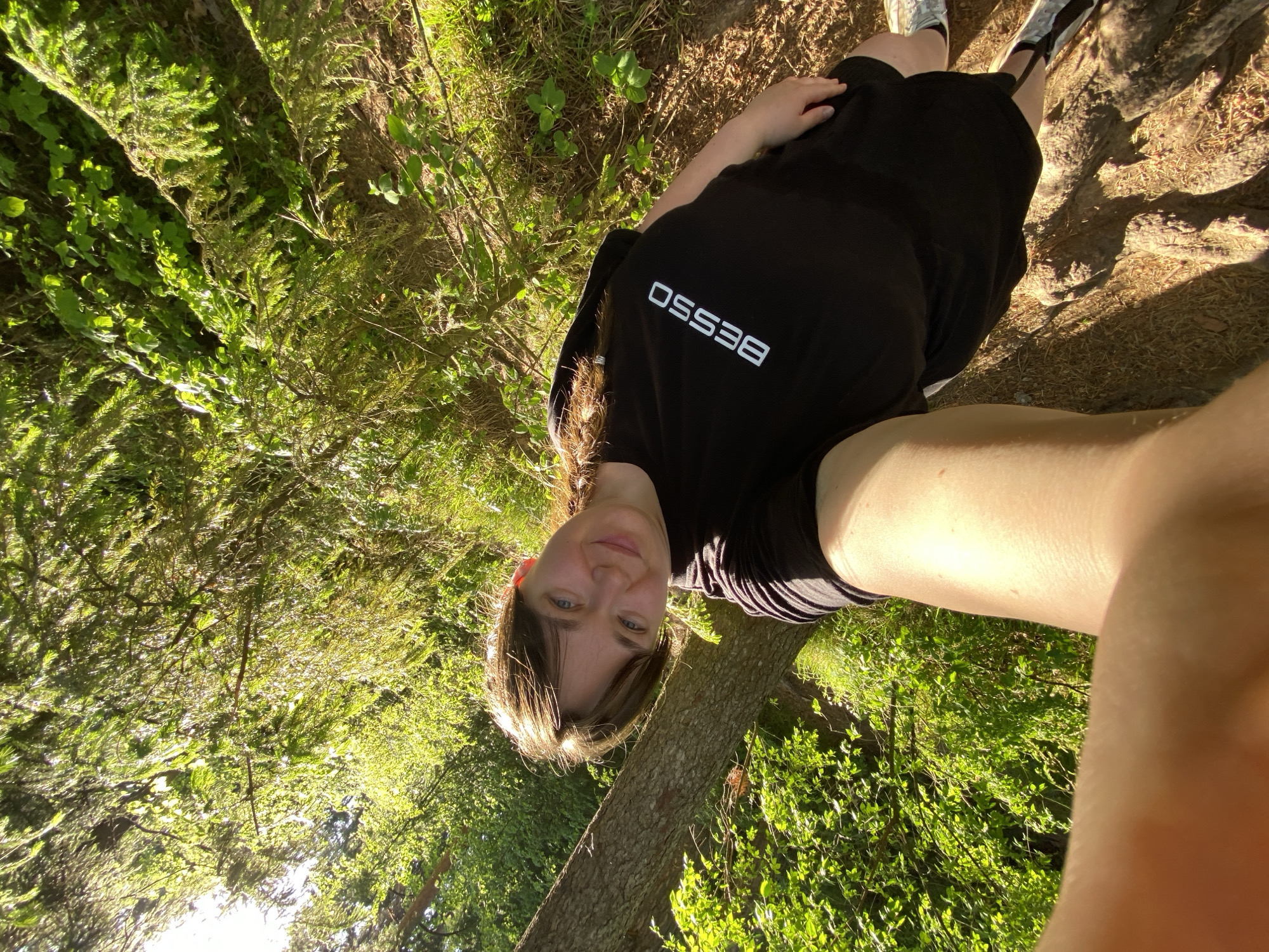 The image is a selfie taken outdoors. A young person is centered, wearing a black t-shirt with "BESSO" printed in white. They are positioned next to a tree trunk, with lush green foliage surrounding them. The environment suggests a park or wooded area, possibly within the city of Votkinsk, Russia. The lighting indicates it is daytime. The subject is looking directly at the camera, and the angle suggests they are holding the camera. The background is filled with trees and vegetation, creating a natural setting.