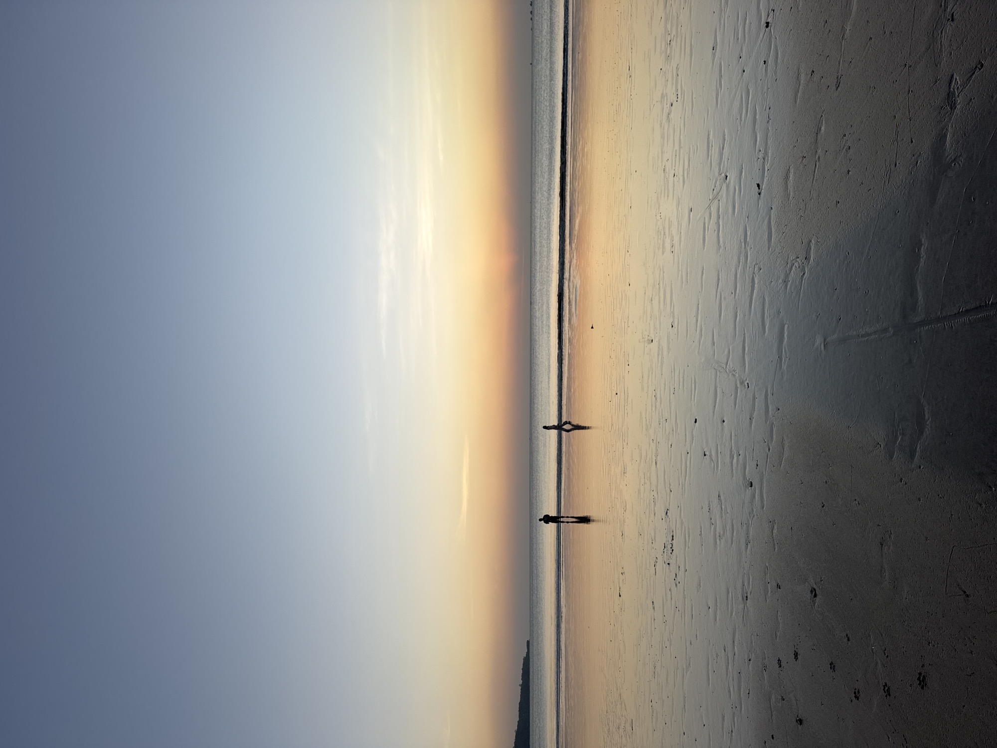 A wide-angle, eye-level photograph captures a serene sunset scene at Kuta Selatan beach in Indonesia. The sky transitions from a soft blue at the top to a warm gradient of orange and yellow near the horizon, with the sun appearing as a bright, diffused glow. The ocean is calm, with a thin line of gentle waves lapping the shore. The wet sand in the foreground reflects the sky's colors, creating a shimmering surface with subtle textures and patterns left by the receding tide. Two figures, silhouetted against the bright horizon, are walking along the water's edge. One figure is slightly closer to the left, facing right, while the other is to the right, facing left. Their reflections are clearly visible in the wet sand. In the distance, a dark, low-lying landmass, possibly a cliff or hills, forms the left edge of the horizon. The overall impression is one of tranquility and natural beauty as day transitions into night.