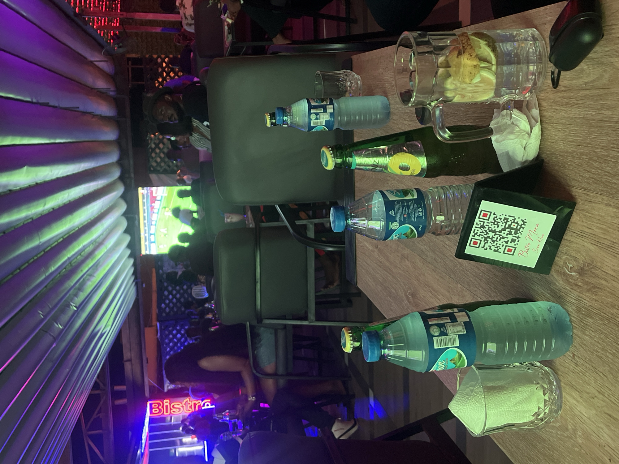 This image depicts an indoor scene within the Boogie Night Club, located in Abuja, Nigeria. The environment is a dimly lit lounge or bar area, illuminated by a dynamic lighting scheme featuring purple and green LED strips integrated into a pleated ceiling structure. A large screen at the rear displays a football (soccer) match. A prominent red neon sign spelling "Bistro" is affixed to a wall on the left.
The foreground is dominated by a wooden table holding various beverages and personal items. Specifically, four plastic bottles of "Safiyah Spring Waters" (50cl) are visible, with two open and partially consumed. Two green glass bottles are present, one clearly labeled "TOUZ Apple." A clear glass mug with a handle contains water infused with multiple lemon or lime slices. Two additional clear glasses are on the table, one containing a folded white paper napkin. A small black stand with a QR code is centrally placed, bearing the text "Bistro Menu Scan Here" and, in script, "Boogie Night Club." A crumpled white paper napkin and a black car key fob or wireless earbud case are also on the table.
Several patrons occupy dark-colored upholstered chairs and booths in the mid-ground and background. A man with a beard, wearing a dark hat and shirt, is seated with his back partially towards the camera, looking to the right. To his left, a woman with long dark hair is seen from behind. Further left, another woman with long dark hair, dressed in a dark top and denim shorts, is seated, engaged with a mobile phone. Other individuals are visible as indistinct figures and silhouettes throughout the club, engaged in socializing and watching the sports broadcast. The floor appears to be a dark, reflective surface.