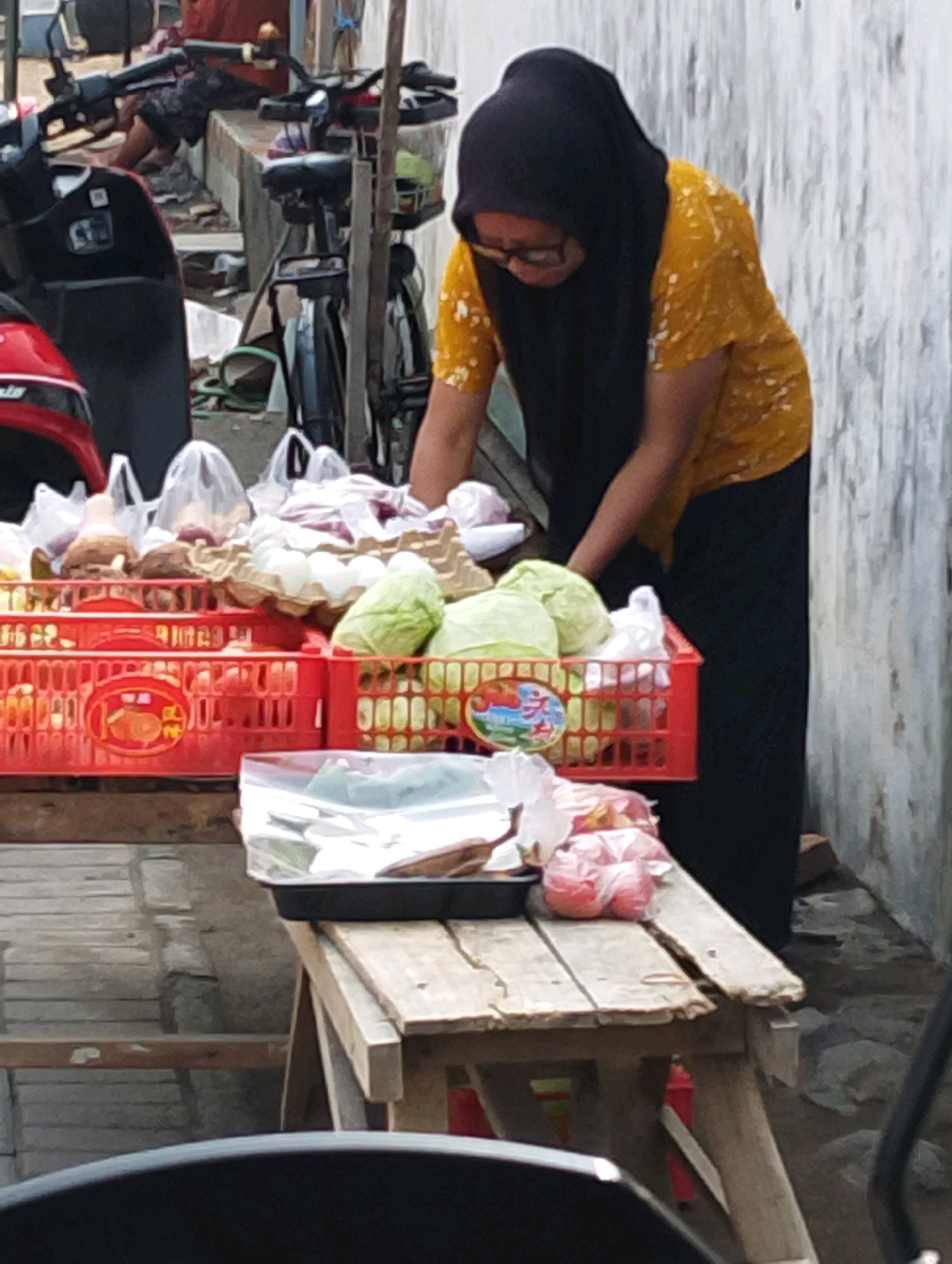 A woman wearing a black hijab and a yellow floral shirt is standing next to a stall selling produce in Glanggang, Indonesia. She is leaning over the stall, which is stocked with red plastic crates containing eggs, cabbage, and other unidentifiable items. Bags of produce are also visible. In the background, there are motorcycles parked and a partially visible building. The scene appears to be outdoors, with a concrete wall and debris on the ground.