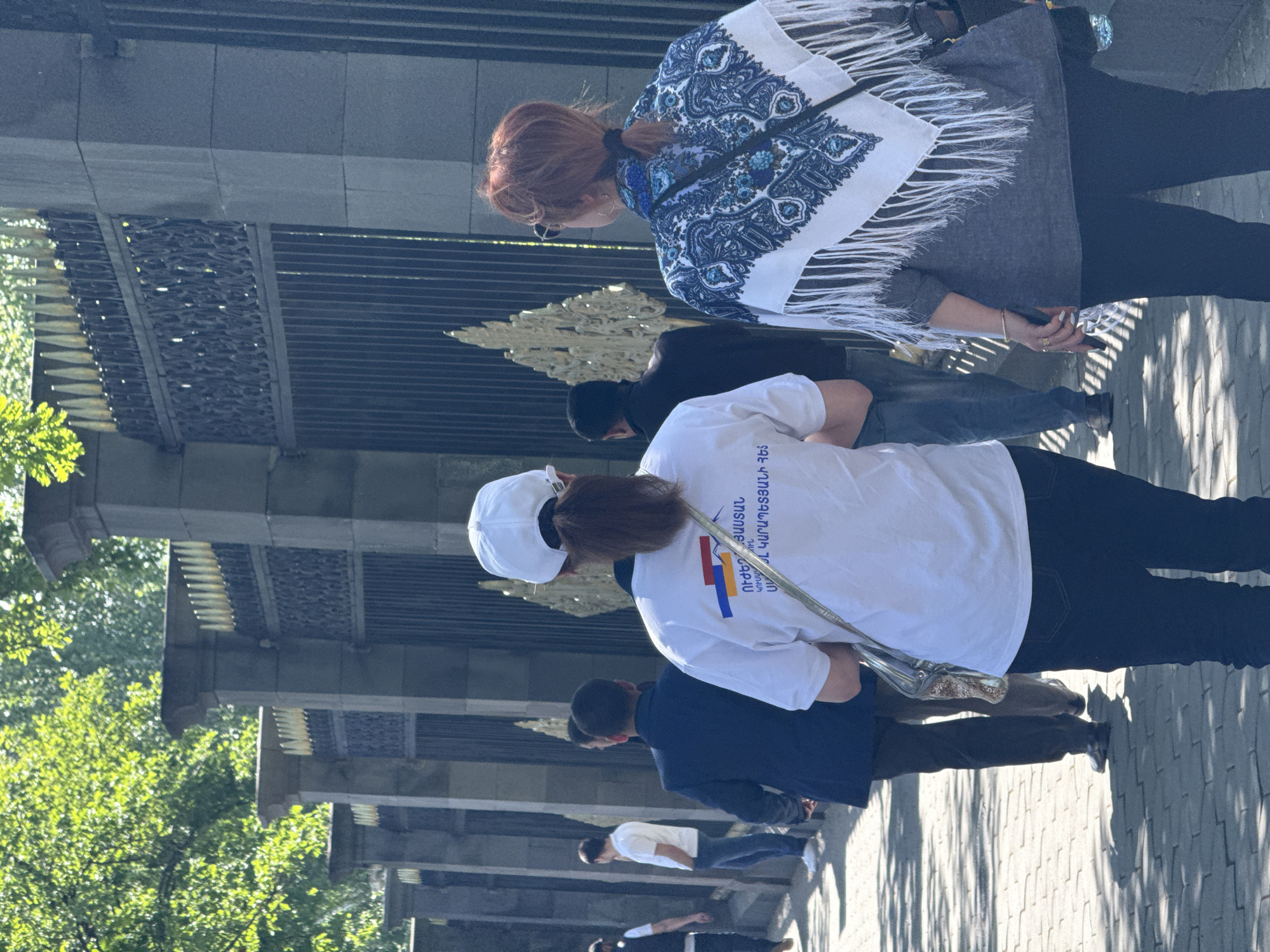 Captured outdoors in Yerevan, Armenia, the scene unfolds along a paved pathway, likely a sidewalk. Pedestrians are present, each with distinctive attire and actions. At the forefront stands a woman clad in a white scarf adorned with blue and white patterns, paired with a grey top and dark trousers. Nearby, another individual sports a white T-shirt featuring the Armenian flag, matched with dark pants and a white baseball cap. The attire suggests a nationalistic pride. Other individuals stroll casually along the pathway, enhancing the sense of an ordinary day in the city. The setting features an ornate metal gate or fence, providing depth to the composition. On the left, lush trees offer a green contrast to the urban architecture, and the sunlight casts shadows that create a dynamic visual effect. The overall impression is that of an unposed, candid capture, indicative of daily life and local expression in Yerevan.
