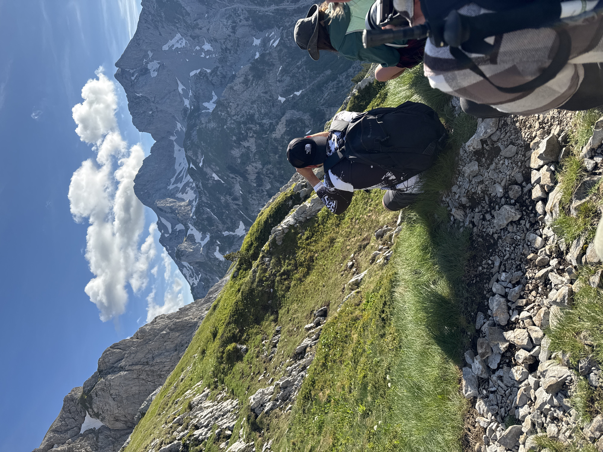 The image depicts a scenic mountain landscape with three individuals seated on a grassy incline, observing the vista. The person in the foreground, with their back to the camera, wears a black baseball cap with a white logo and a black backpack. To their right, another person, also facing away, is wearing a green jacket and a wide-brimmed hat. A third person's shoulders and backpack are visible on the far right. The background is dominated by rugged, snow-capped mountains under a bright blue sky with scattered clouds. The terrain is rocky and covered with green grass and sparse vegetation. This scene is likely located in the Durmitor National Park in Montenegro, a region known for its dramatic mountain peaks and natural beauty.