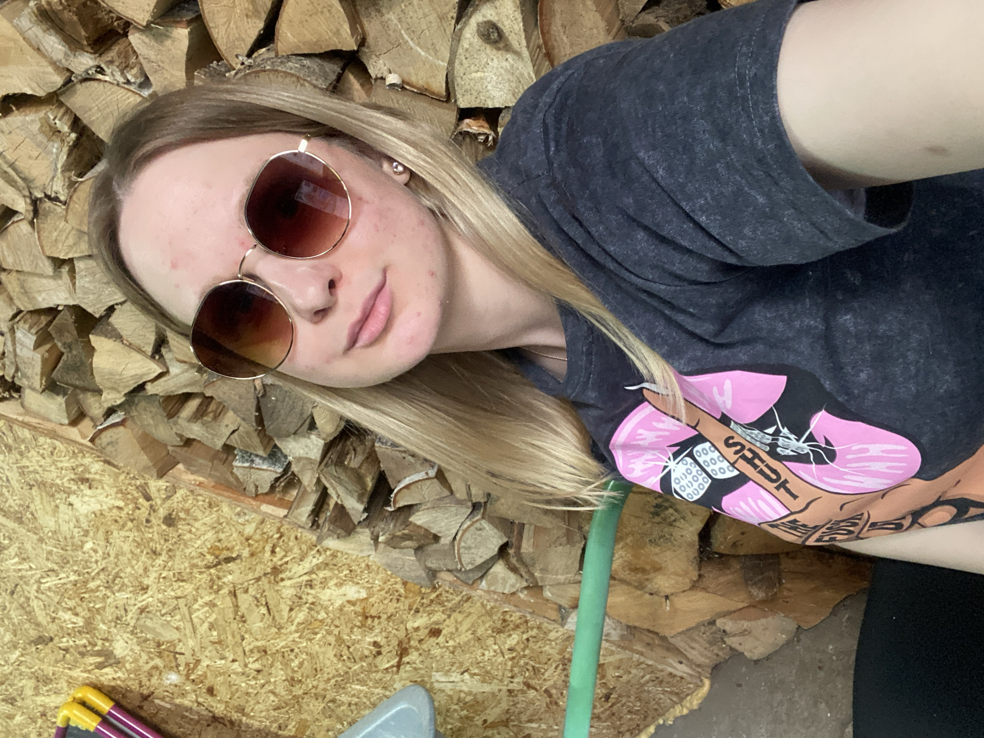 A young adult female is depicted in a medium close-up, selfie-style photograph. She has long, straight blonde hair and is wearing large, round sunglasses with brown-tinted lenses and thin, gold-toned metal frames. Her complexion displays multiple red blemishes across her forehead, cheeks, and chin. A small, clear or white stud earring is visible in her right earlobe. She wears a dark gray, short-sleeved t-shirt with a textured, possibly distressed, fabric. The t-shirt features a prominent graphic on the left chest/shoulder area, depicting an anthropomorphic, light-brown colored hand with text reading "SHUT THE FUCK UP" across the fingers, positioned against a large, pink, floral or starburst design.
The background consists of two distinct elements: to the viewer's left, a wall constructed of oriented strand board (OSB), and to the right, a densely stacked pile of split firewood, appearing to be stored indoors. On the OSB wall, several objects are partially visible: a light gray curved plastic item in the upper left, a yellow-handled tool, and a purple-handled tool hanging on the wall. A green hose or pipe runs horizontally across the lower-left quadrant of the image. The overall setting appears to be an interior space, likely a utility area, garage, or wood storage facility in Unknown, Unknown.