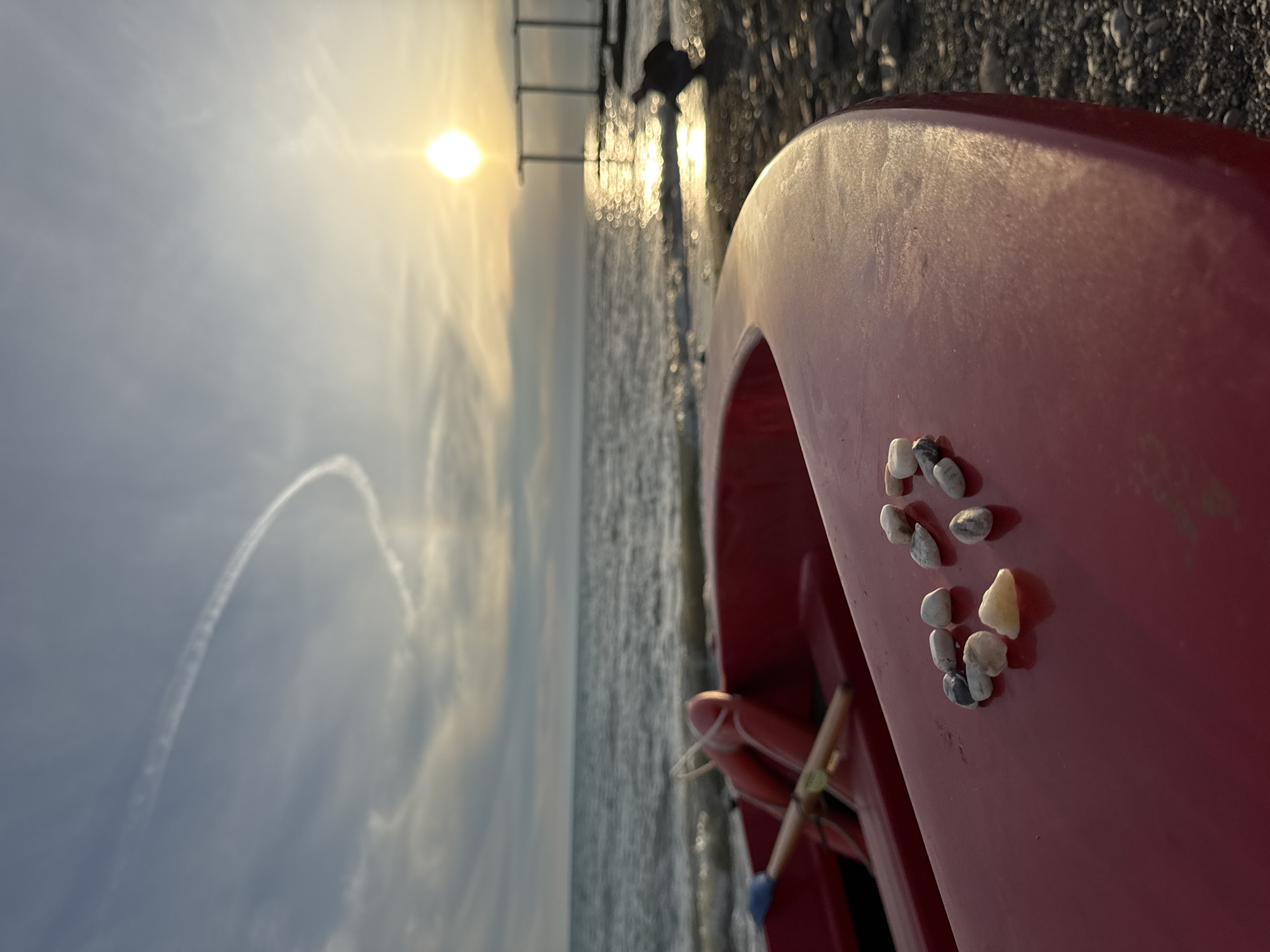 A red boat rests on a pebble beach, its bow close to the viewer, with a cluster of small, smooth stones arranged on its surface. The sea stretches out beyond the boat, calm with gentle waves reflecting the golden sunlight of a setting sun. In the distance, a pier with several vertical supports juts into the water. A faint contrail forms a curved line in the pale blue sky, and a solitary figure is visible in the middle ground, partially obscured by glare from the water. The scene appears to be located on the coast of Sochi, Russia, given the architecture of the pier and the overall atmosphere.