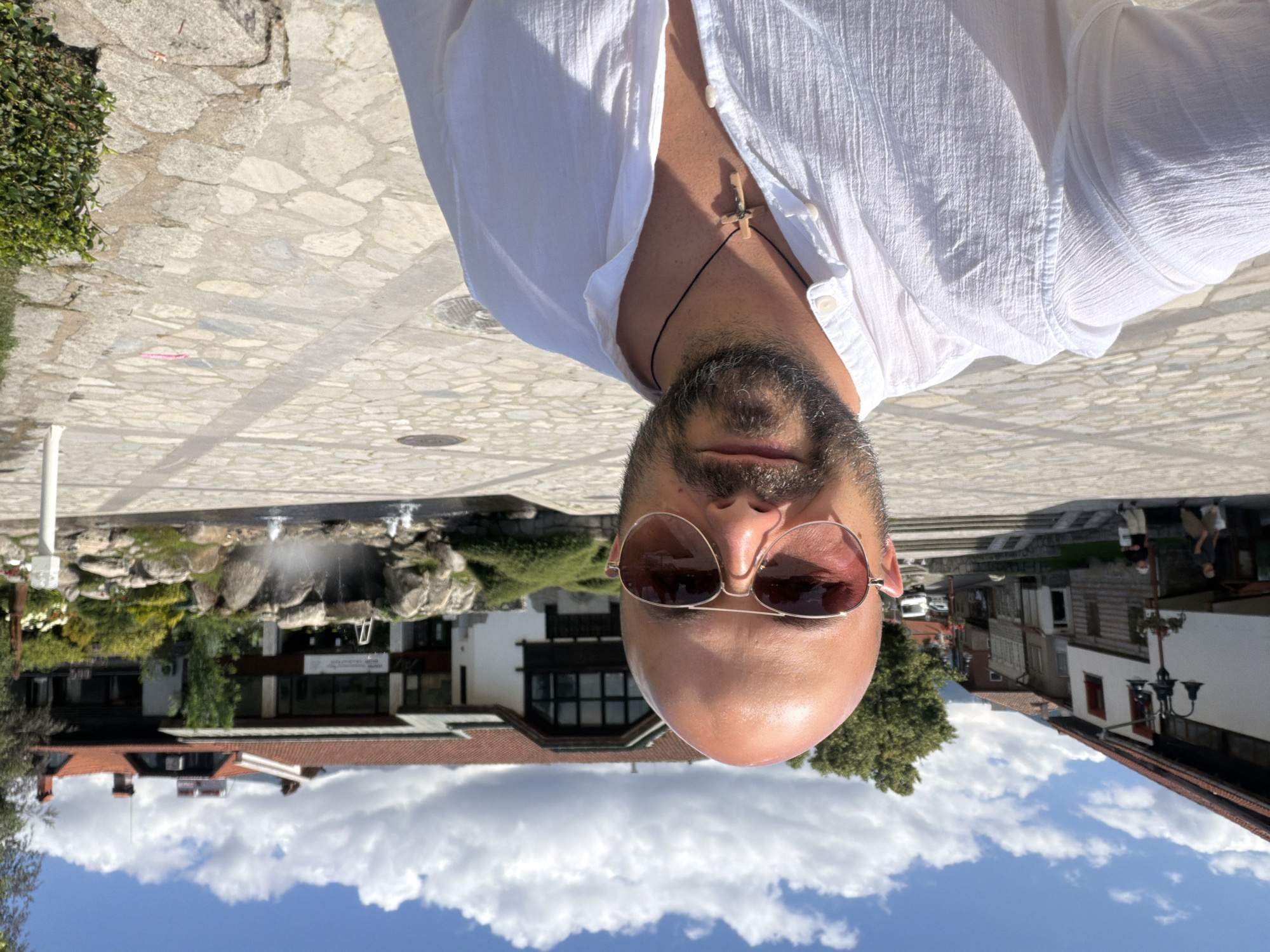 A bald, bearded man wearing sunglasses and a white open-collared shirt with a wooden cross necklace is taking a selfie in a cobblestone plaza in Bansko, Bulgaria. Behind him, a modern building with a red-tiled roof features a waterfall and a decorative rock garden. In the background, traditional Bulgarian houses line a street under a partly cloudy blue sky.