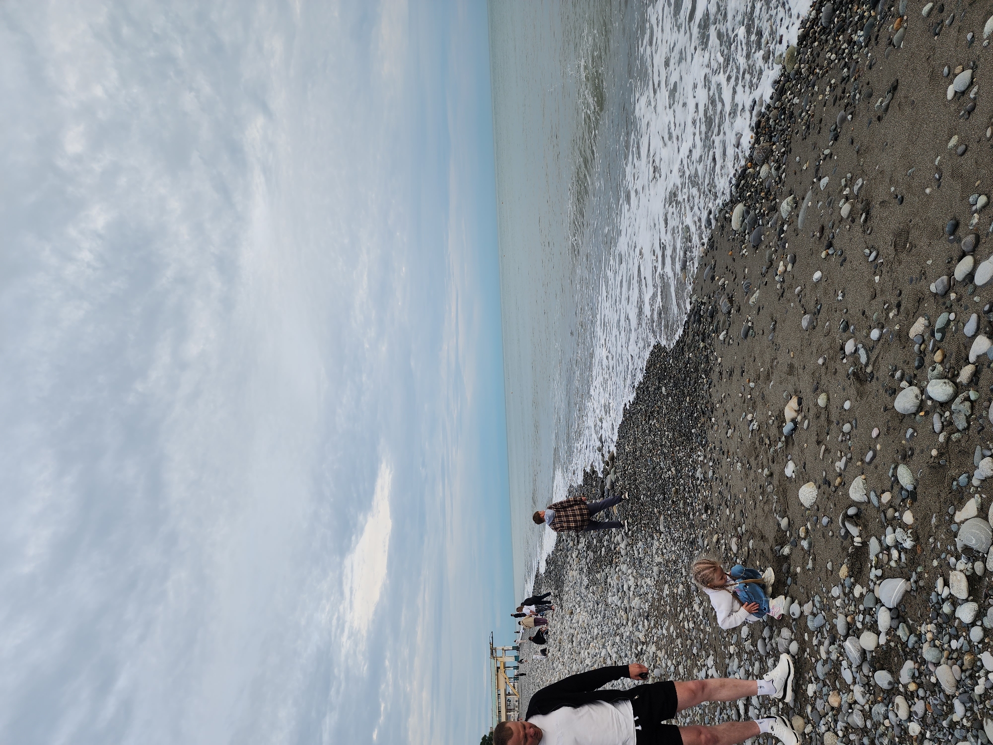 The image depicts a pebble beach scene in Sochi, Russia, under an overcast sky. The horizon line is visible, separating a muted blue-grey sky with scattered clouds from the calm, greyish-blue sea. Gentle white waves are breaking along the shoreline, contrasting with the dark, wet pebbles.
In the foreground, a person's legs and torso are visible, wearing a white t-shirt, black shorts, and white sneakers with black socks. To their right, a young child, likely a girl with blonde hair in braids, is sitting on the pebble beach, facing towards the water. She is dressed in a white long-sleeved top and blue jeans, appearing to be observing or playing with the pebbles.
Further along the beach, closer to the water, another individual wearing a plaid shirt and dark pants stands, looking out at the sea. Several other people are present in the mid-ground, casually walking or standing on the extensive pebble and sand beach. In the distance, a light-colored structure, possibly a pier or platform, extends into the sea. The beach comprises a mix of various sized grey, white, and darker pebbles interspersed with patches of darker sand. The lighting is soft and diffused, indicating an overcast or cloudy daytime. No visible text is discernible in the image.