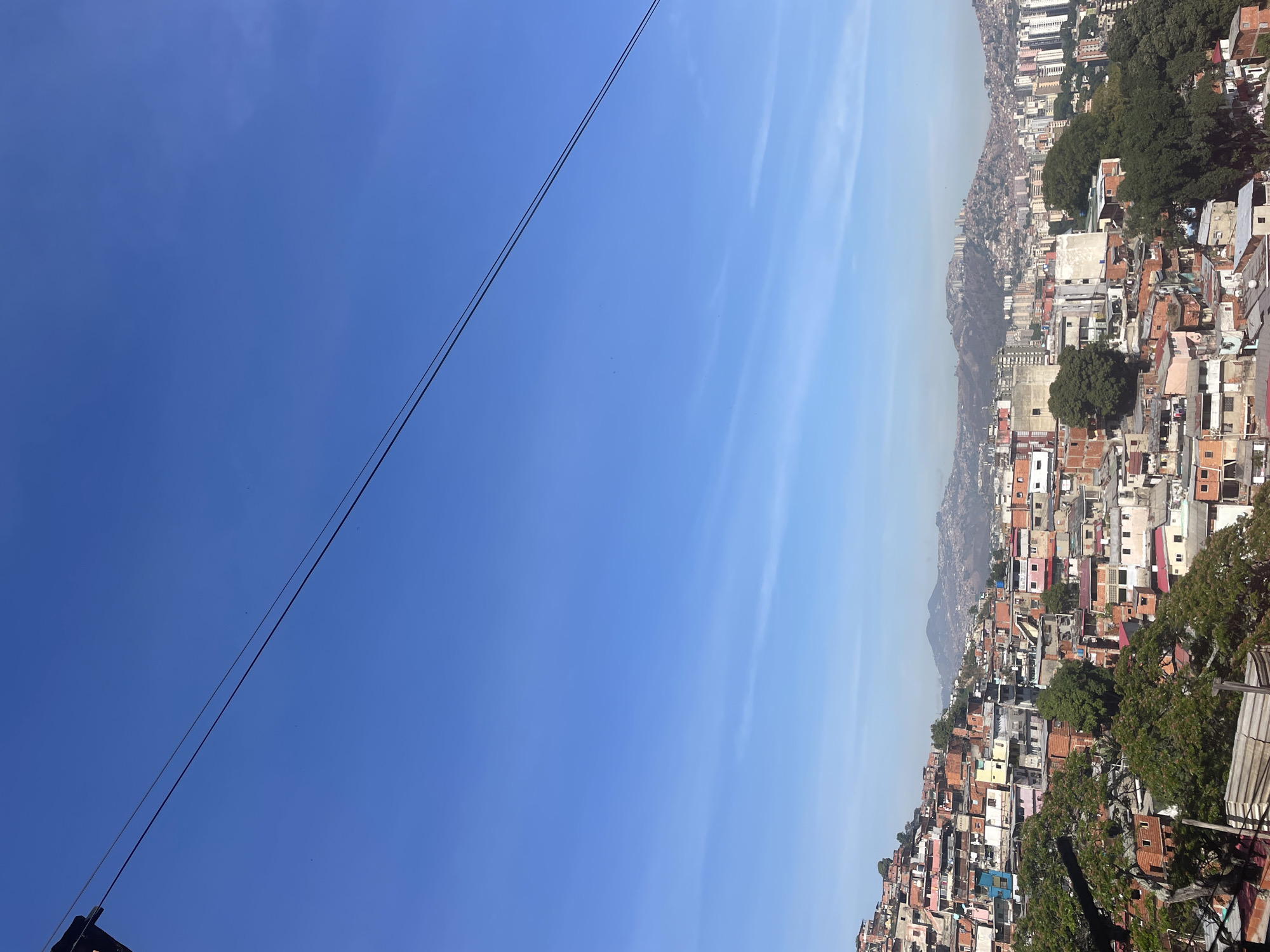 The image presents a view of a densely populated area, a favela, in Caracas, Venezuela, under a clear blue sky. The cityscape is characterized by a patchwork of closely built houses, varying in colors and construction materials, stretching up a hillside. In the background, taller buildings emerge, suggesting the proximity of more urbanized areas. Overhead, a set of power lines cut across the top left corner of the frame.