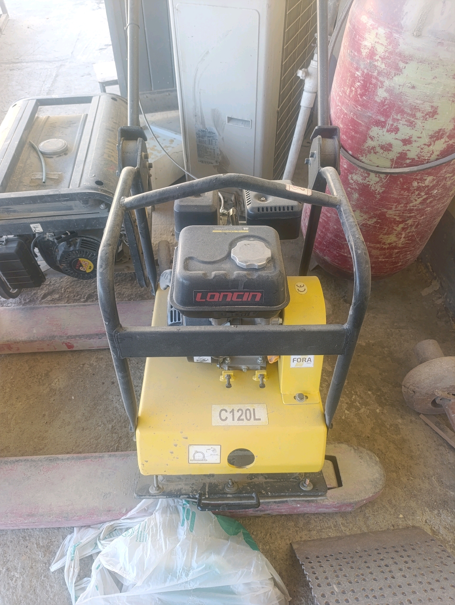 The image depicts a construction site in Navoiy City, Uzbekistan, focusing on a yellow vibratory plate compactor labeled "C120L" and "LONCIN". The compactor is positioned on a dusty ground, with a red and white cylindrical object, possibly a fuel tank, to its right. In the background, a white air conditioning unit is visible. To the left of the compactor, a black portable generator is partially obscured. A white plastic bag lies on the ground in the foreground. The overall scene suggests an active or recently used work area, characterized by industrial equipment and a dusty environment, typical of construction zones in an urban setting.