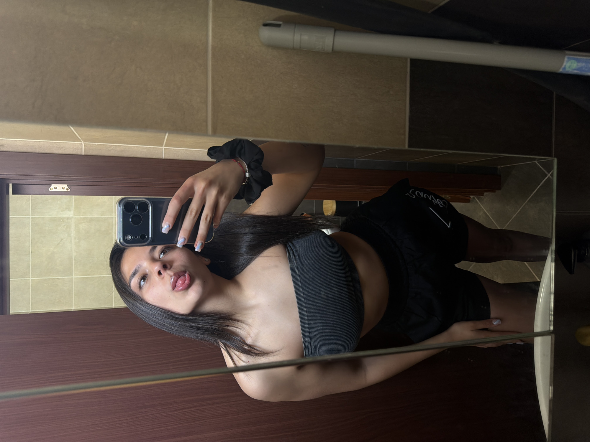 A young woman is captured in a mirror selfie, showing her reflection from the chest up. She is looking at her smartphone, which she holds in her right hand, and is playfully sticking out her tongue with her lips slightly parted. Her dark hair is long and falls over her shoulders. She is wearing a black ribbed bandeau top and black shorts with white, mirrored text that appears to spell "SAVAGES" on the left thigh. On her left wrist, she wears a black scrunchie and a thin bracelet with a small charm. Her nails are painted with a light colored polish, possibly white or light blue, with subtle designs.
The setting appears to be an indoor space, likely a bathroom or dressing area, based on the tiled walls and mirror. The upper part of the wall seen in the reflection is covered with light brown rectangular tiles, separated by lighter grout lines. Below this, there is a brown wooden panel or frame. Further into the background, darker brown and black tiles are visible. A white pipe or conduit runs horizontally across the upper background. The mirror itself has a thin golden or metallic frame visible along its edge.
The activity is a casual mirror selfie, with the woman expressing a playful mood. No specific time of day or weather is discernible as it's an indoor shot. The location context is Gabrovo, Bulgaria.