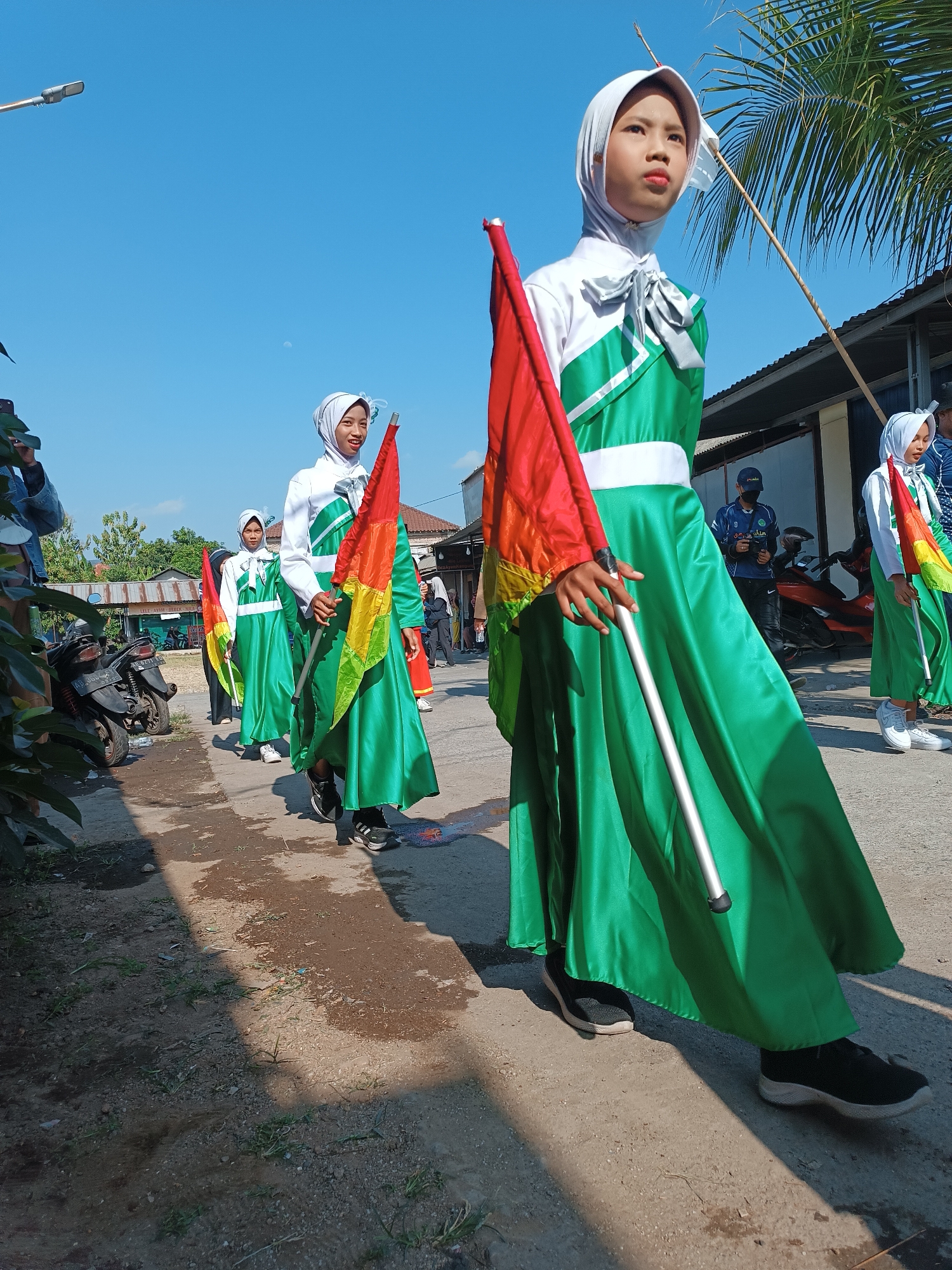 In Imogiri, Indonesia, a group of young girls in matching green and white uniforms, complete with white hijabs, are participating in a parade. The uniforms feature a white top with green accents and a white belt, paired with a long, flowing green skirt. Each girl holds a flag pole, with one girl prominently displaying a flag that transitions from red to yellow to green. The girls are walking along a paved road, with the foreground showing a dirt and gravel area. In the background, there are buildings, a parked motorcycle, and trees, including a large palm tree. The sky is clear and blue, suggesting a sunny day. A street lamp is visible in the upper left corner of the image. The overall scene depicts a community event or celebration involving youth.
