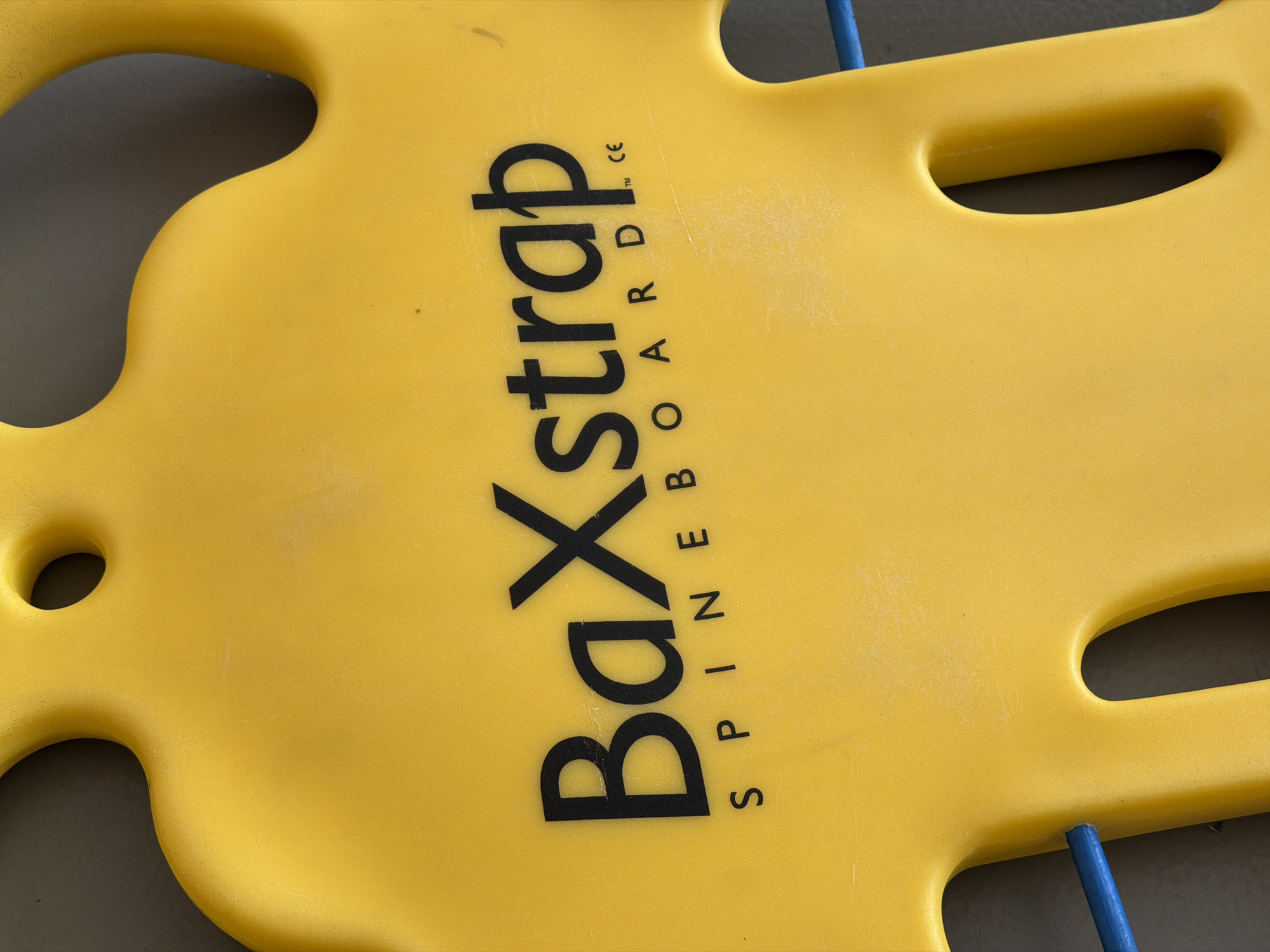 The image captures a close-up view of a yellow medical spineboard. The board is made of bright yellow plastic and shows signs of wear, including minor scratches and scuffs across its surface. It features several large, irregularly shaped cutouts, likely designed as handholds or attachment points for straps.
The most prominent feature is the black text printed diagonally across the board. The main text reads "BaXstrap" in large, bold letters. Below this, in smaller, vertically oriented lettering, the word "SPINEBOARD" is visible. Adjacent to the "p" in "BaXstrap", small symbols "™ CE" are also present.
Through the cutouts, a dark, undifferentiated background is visible. Small blue elements, possibly parts of securing straps or structural components of the board, are visible near the top and bottom edges of the frame. No people or specific activities are depicted; the spineboard is stationary. The lighting is even and diffused, highlighting the texture of the plastic. No specific location cues are discernible from the image itself.