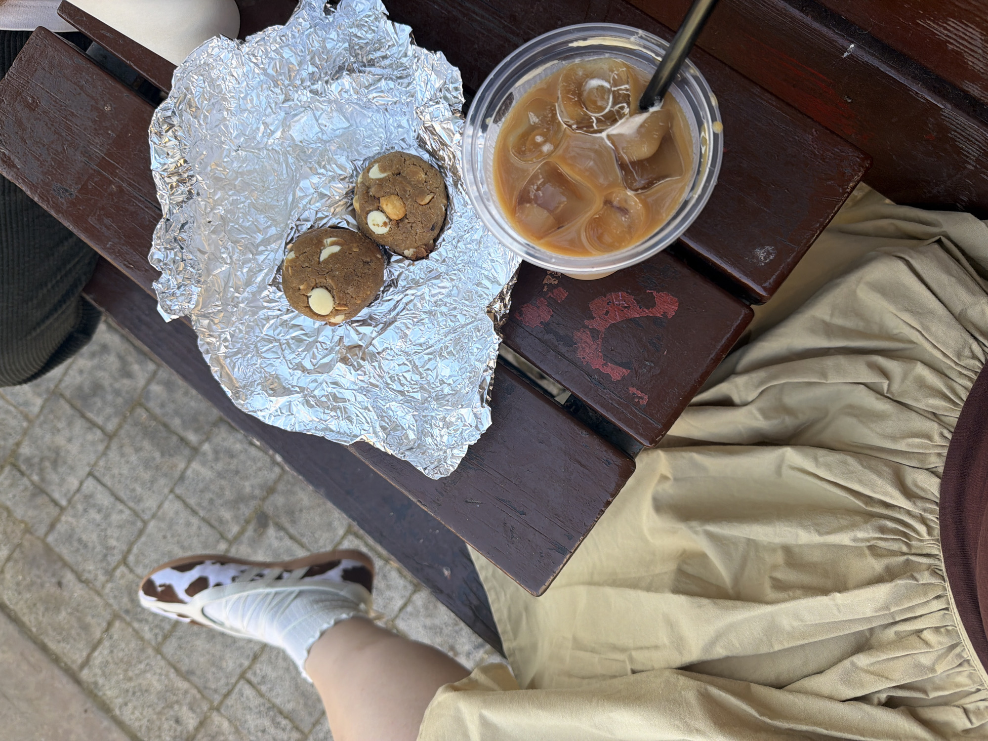 On an outdoor wooden bench in Tel Aviv, Israel, a person is enjoying a snack and a drink. Two round, brown cookies with white chocolate chips sit on a crumpled piece of aluminum foil. Next to the cookies is a clear plastic cup filled with a light brown iced coffee drink with a black straw. The person's leg is visible, clad in a white sock and a brown-and-white patterned shoe. They are wearing a long, light-beige skirt with a brown trim. The bench is positioned on a paved surface.