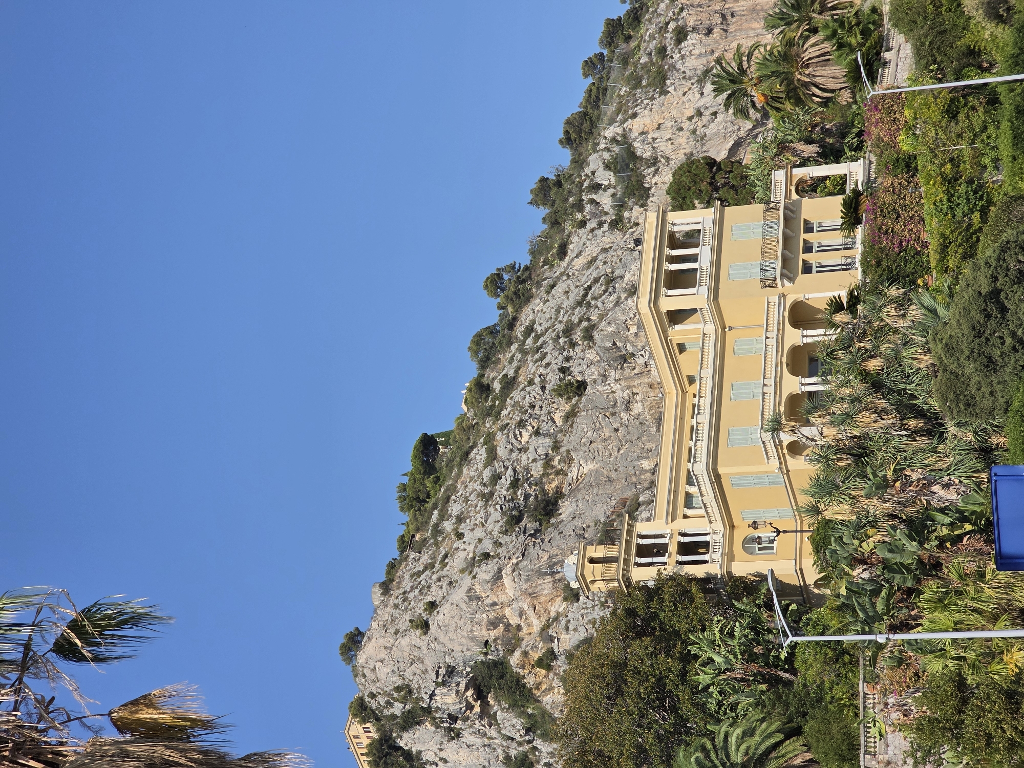 The image depicts a large, pale yellow villa situated on a steep, rocky hillside in Menton, France. The villa has multiple levels, a prominent tower with a dome, and classical architectural elements such as columns and balconies. It is surrounded by lush Mediterranean vegetation, including palm trees, banana trees, and flowering bushes, which cascade down the hillside. The clear blue sky dominates the upper portion of the image, with a sliver of a distant building visible at the top of the hill to the left. A modern white street lamp stands in the foreground, partially obscuring some of the vegetation. The scene is brightly lit, suggesting it was taken during the daytime.