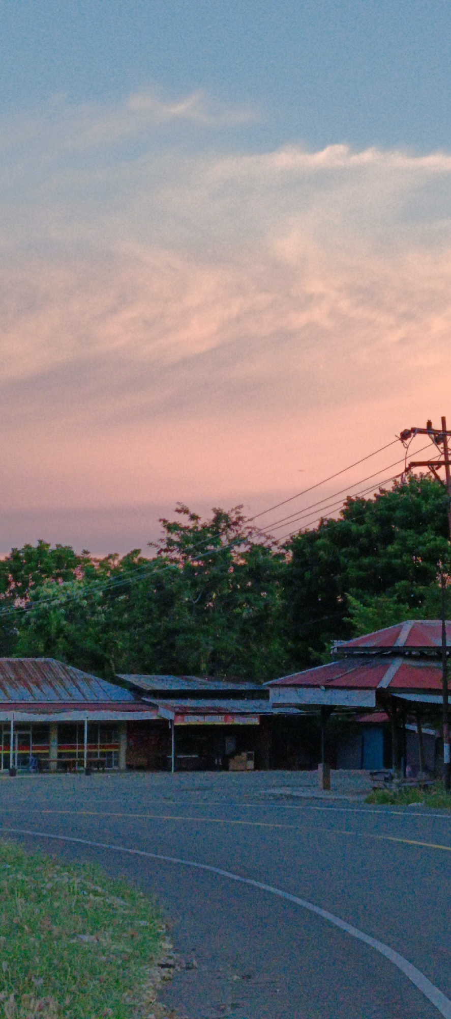 The image depicts a street scene in Sukamulia, Indonesia, at dusk. The sky is a gradient of pale blue and soft pink, with wispy clouds. In the foreground, a paved road curves to the right, with a strip of green grass and vegetation along its left edge. Across the road, several low-lying buildings with corrugated metal roofs are visible. These structures appear to be small shops or stalls. One building has a sign that is partially obscured, but the word "WARUNG" can be discerned, indicating a small general store. Trees with dark green foliage line the background, and a utility pole with several wires stretches across the upper right portion of the image. There are no people or vehicles present in the scene, and the overall atmosphere is quiet and still, indicative of the approaching night.