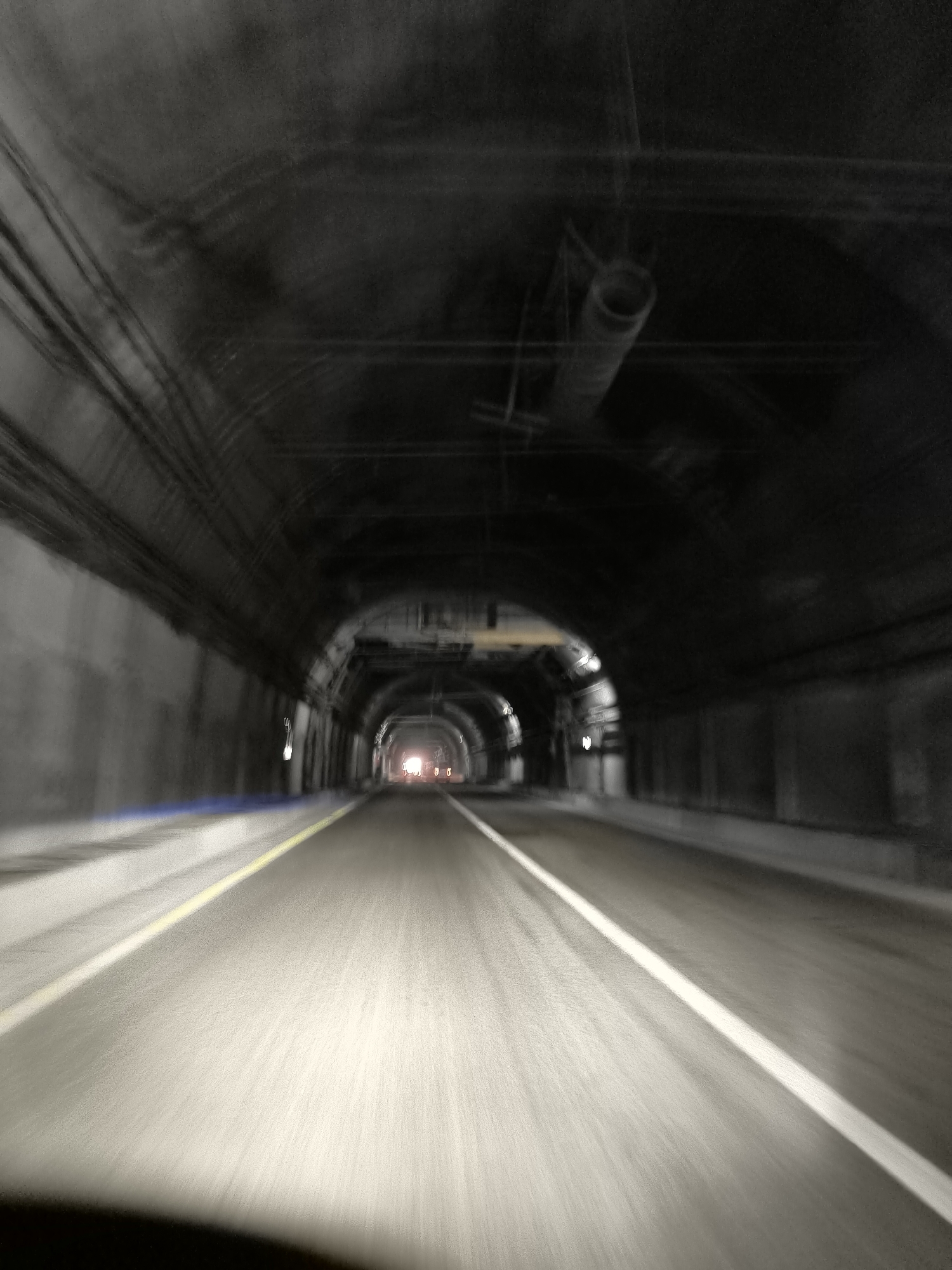 The image depicts the interior of a tunnel, likely a road tunnel, with a sense of motion blur indicating movement. The tunnel appears to be constructed of concrete, with visible infrastructure such as pipes and cables along the ceiling. The road surface is visible in the foreground, marked with white and yellow lines, suggesting lanes of traffic. Lights are visible at the far end of the tunnel, indicating an exit or a section with different lighting. The overall lighting is dim, with illumination coming from the road and distant lights. The environment is dark and enclosed. There are no visible people or specific objects that would identify the location within the city of Sarydala, Uzbekistan.
