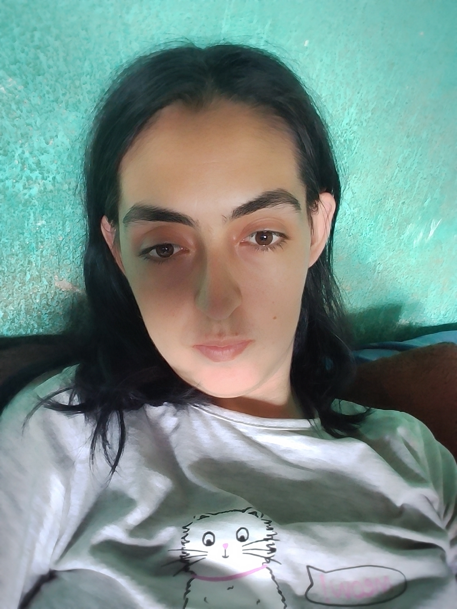 A medium shot portrait shows a young woman with long, dark hair, dark eyebrows, and brown eyes. She is wearing a light-colored t-shirt with a cartoon cat graphic. The background appears to be a textured, turquoise-colored wall. The lighting is dim, casting shadows on the woman's face. The image was taken indoors. No specific location in Stara Zagora, Bulgaria can be identified from the provided image.